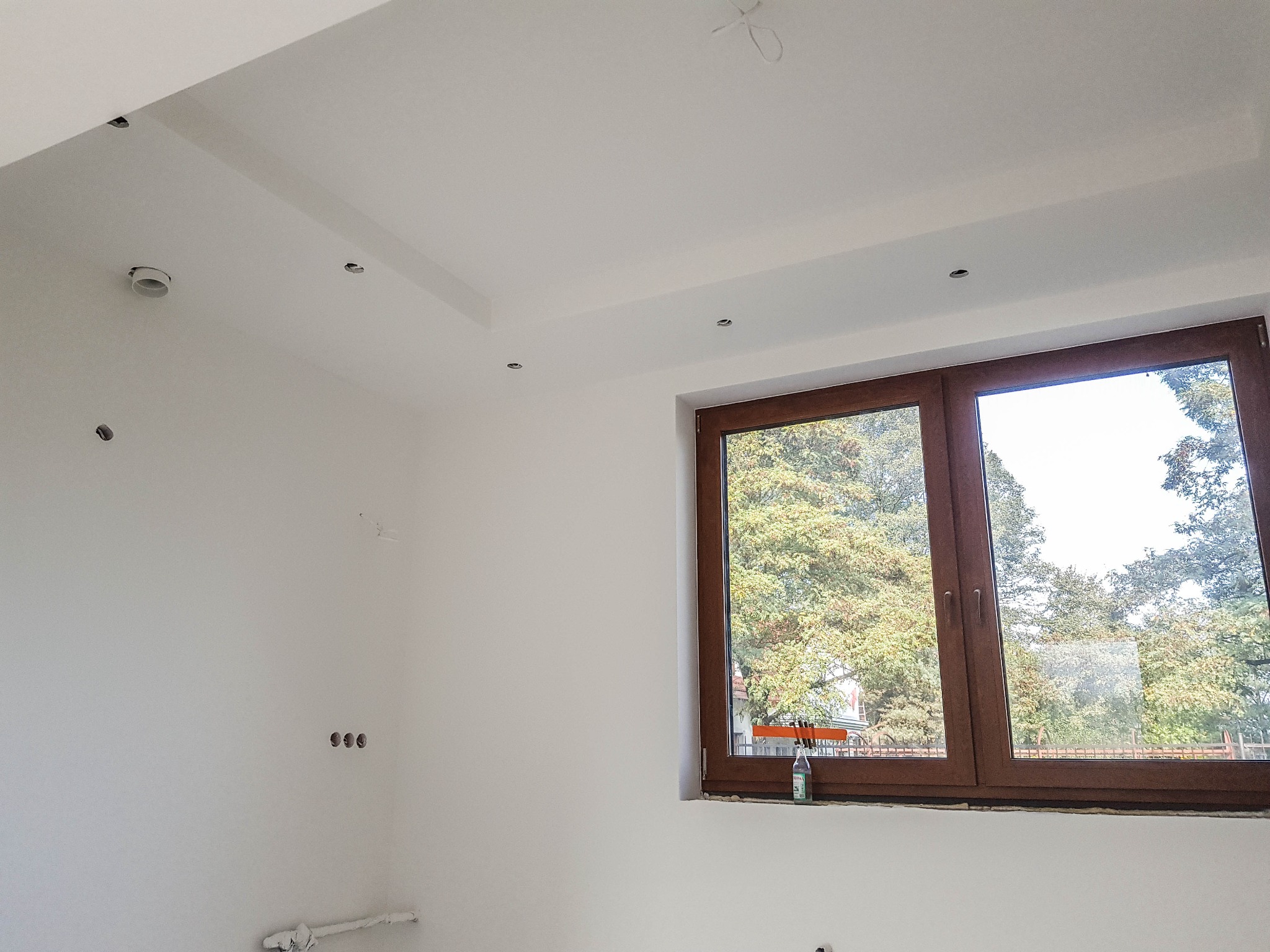 Wnętrze po świeżym pomalowaniu w Czechowice-Dziedzice, widoczne gładkie, białe ściany z przygotowanymi otworami na oświetlenie sufitowe, brązowe okno z widokiem na zieleń, butelka wody mineralnej...