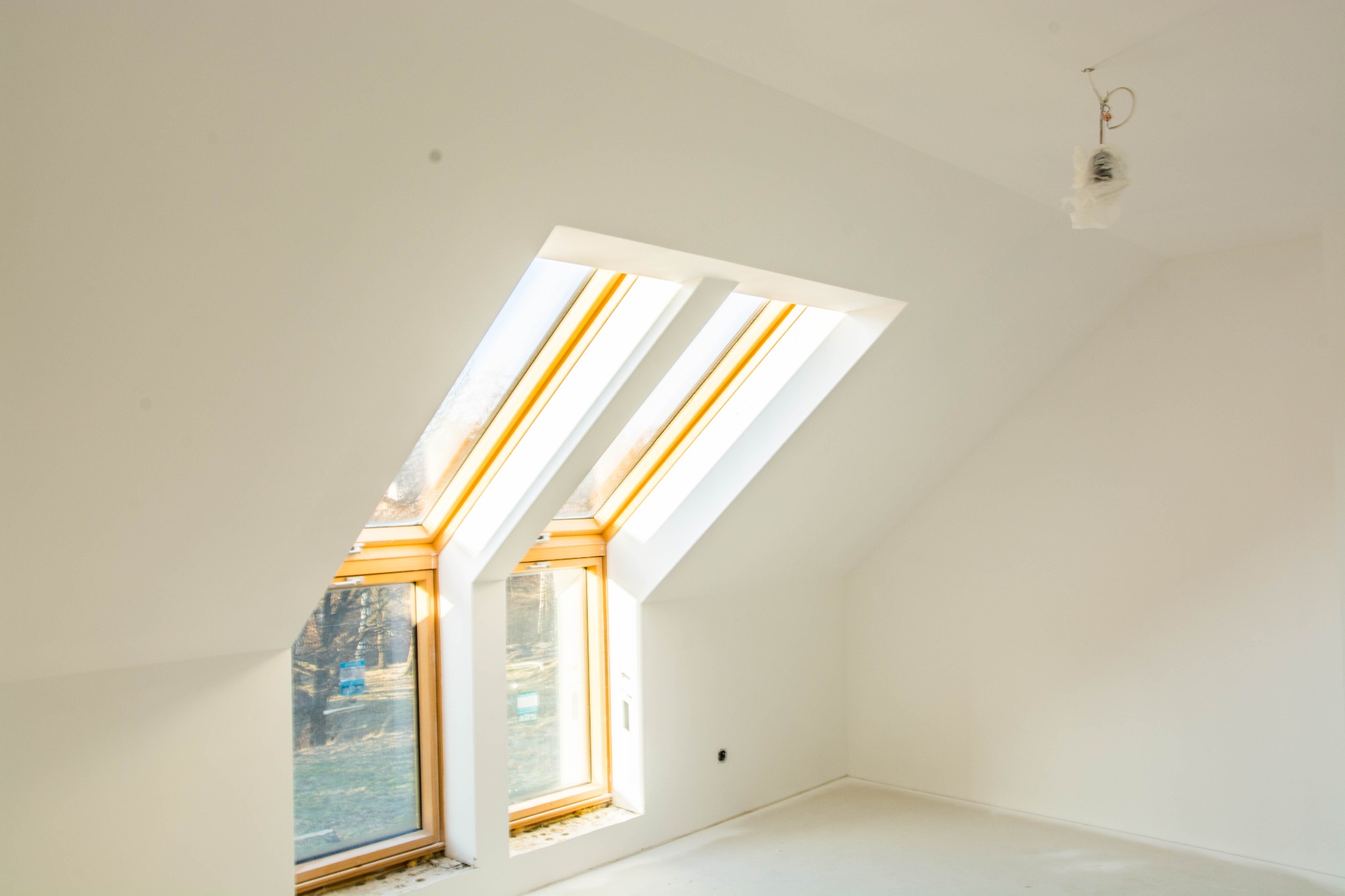 Poddasze po remoncie ze świeżo pomalowanymi ścianami na biało, widoczne okna dachowe z drewnianą ramą i przygotowane miejsce na lampę sufitową.