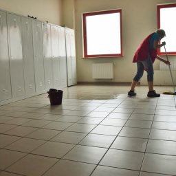 GLAart Katarzyna Glapa - Osoba w czerwonej kamizelce myje podłogę mopem w pomieszczeniu z szarymi szafkami i dwoma oknami, wiadro stoi obok kałuży.