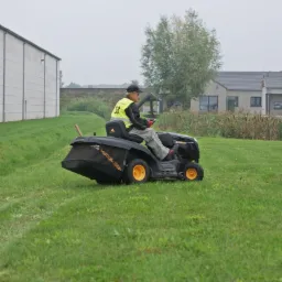 Operator w kamizelce odblaskowej kosi trawę kosiarką samojezdną McCulloch na zielonym trawniku, w tle budynek gospodarczy i dom jednorodzinny.