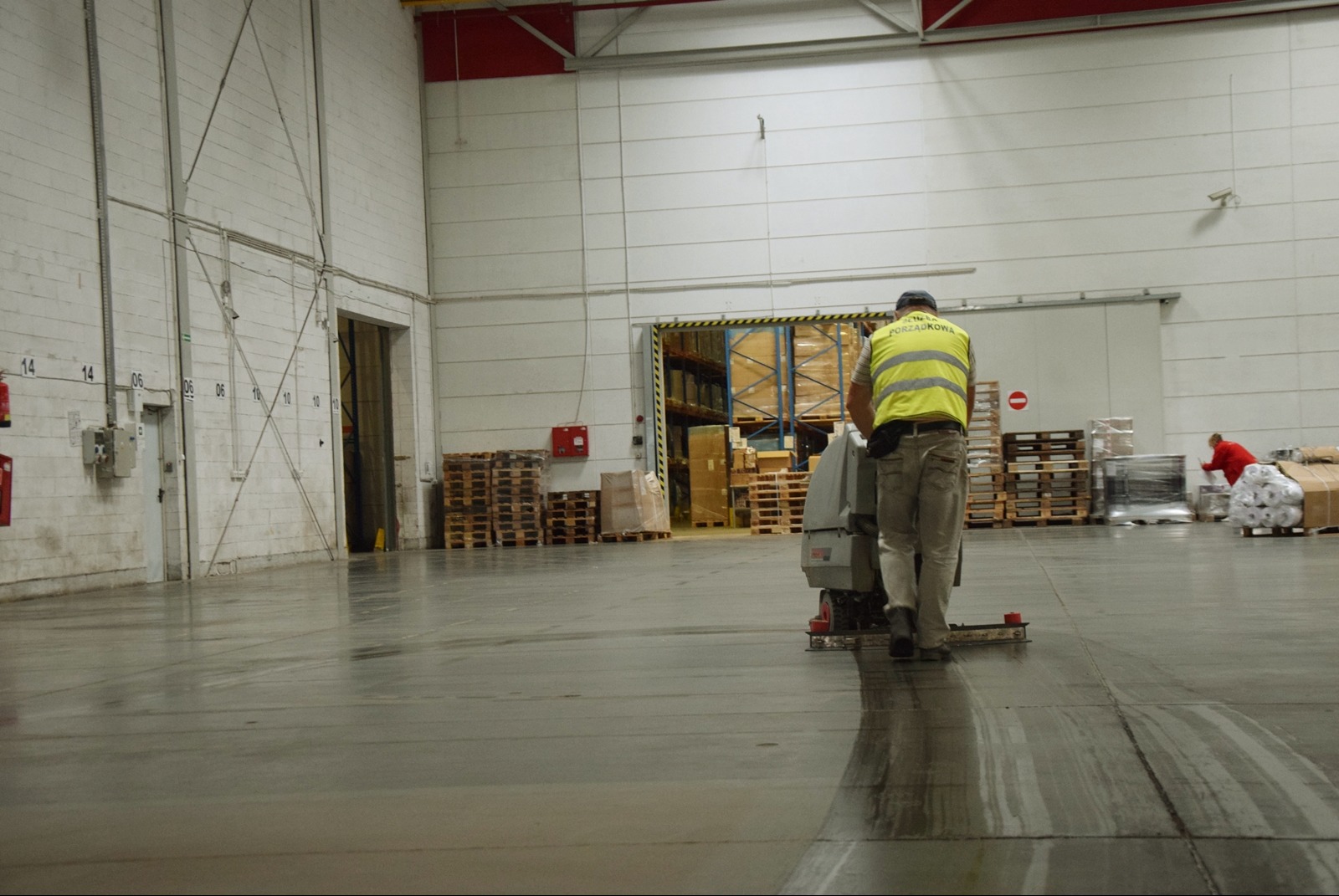 Pracownik w kamizelce odblaskowej czyści posadzkę przemysłową za pomocą specjalistycznej maszyny w przestronnym magazynie z paletami i regałami.