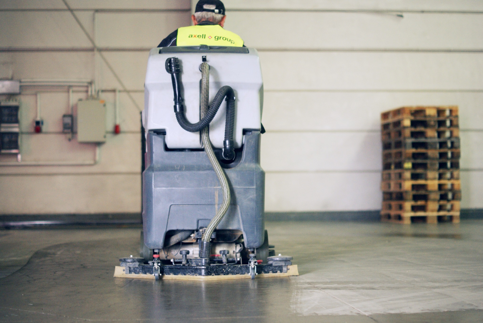 Pracownik obsługuje przemysłową maszynę czyszczącą podłogę w hali magazynowej, widoczny stos palet w tle.