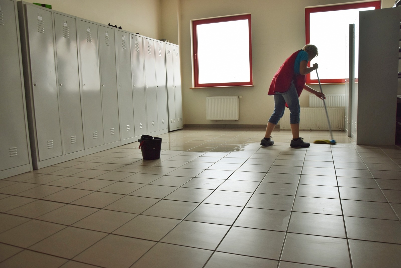 Osoba w czerwonej kamizelce myje podłogę mopem w pomieszczeniu z szarymi szafkami i dwoma oknami, wiadro stoi obok kałuży.