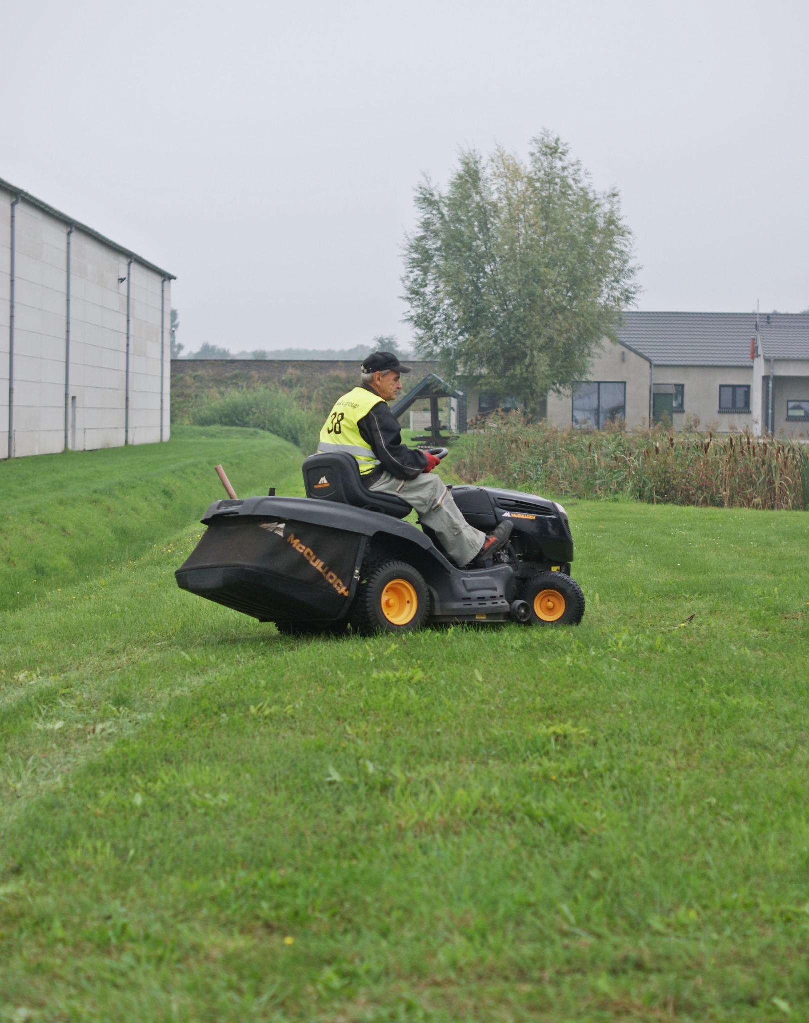 Operator w kamizelce odblaskowej kosi trawę kosiarką samojezdną McCulloch na zielonym trawniku, w tle budynek gospodarczy i dom jednorodzinny.