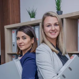 Dwie uśmiechnięte kobiety, brunetka i blondynka, stoją plecami do siebie, trzymając szare segregatory na dokumenty na tle półki z segregatorami i roślinami doniczkowymi.