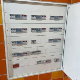 ARKON Electric - Rozdzielnia elektryczna z licznikami, zamontowana na ścianie wyłożonej pomarańczowo-żółtymi płytkami. Drzwiczki szafki otwarte, widoczne zabezpieczenia i elementy instalacji.