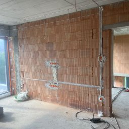 ARKON Electric - Instalacja elektryczna w surowym budynku: przewody w peszlach, puszki podtynkowe w ścianie z cegły dziurawki, widoczne kable na suficie i podłodze. Etap prac.