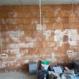 ARKON Electric - Surowa ściana z cegły z puszkami instalacyjnymi i przewodami. Widoczne narzędzia i materiały budowlane. Stan surowy przed tynkowaniem. Prace elektryczne w trakcie.