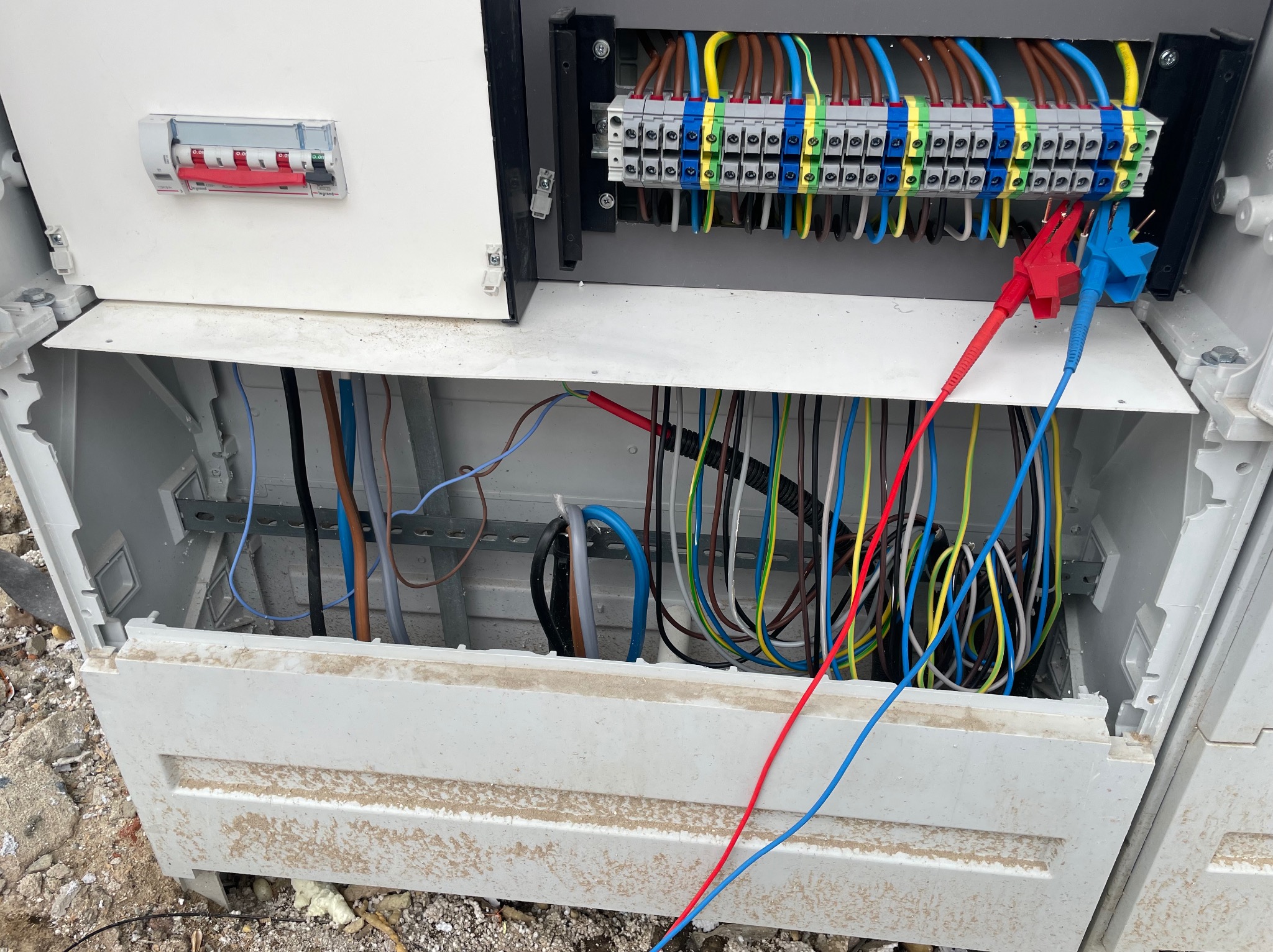 Otwarta skrzynka elektryczna z podłączonymi przewodami pomiarowymi (czerwony i niebieski), widoczne bezpieczniki i listwa zaciskowa, obudowa ubrudzona.