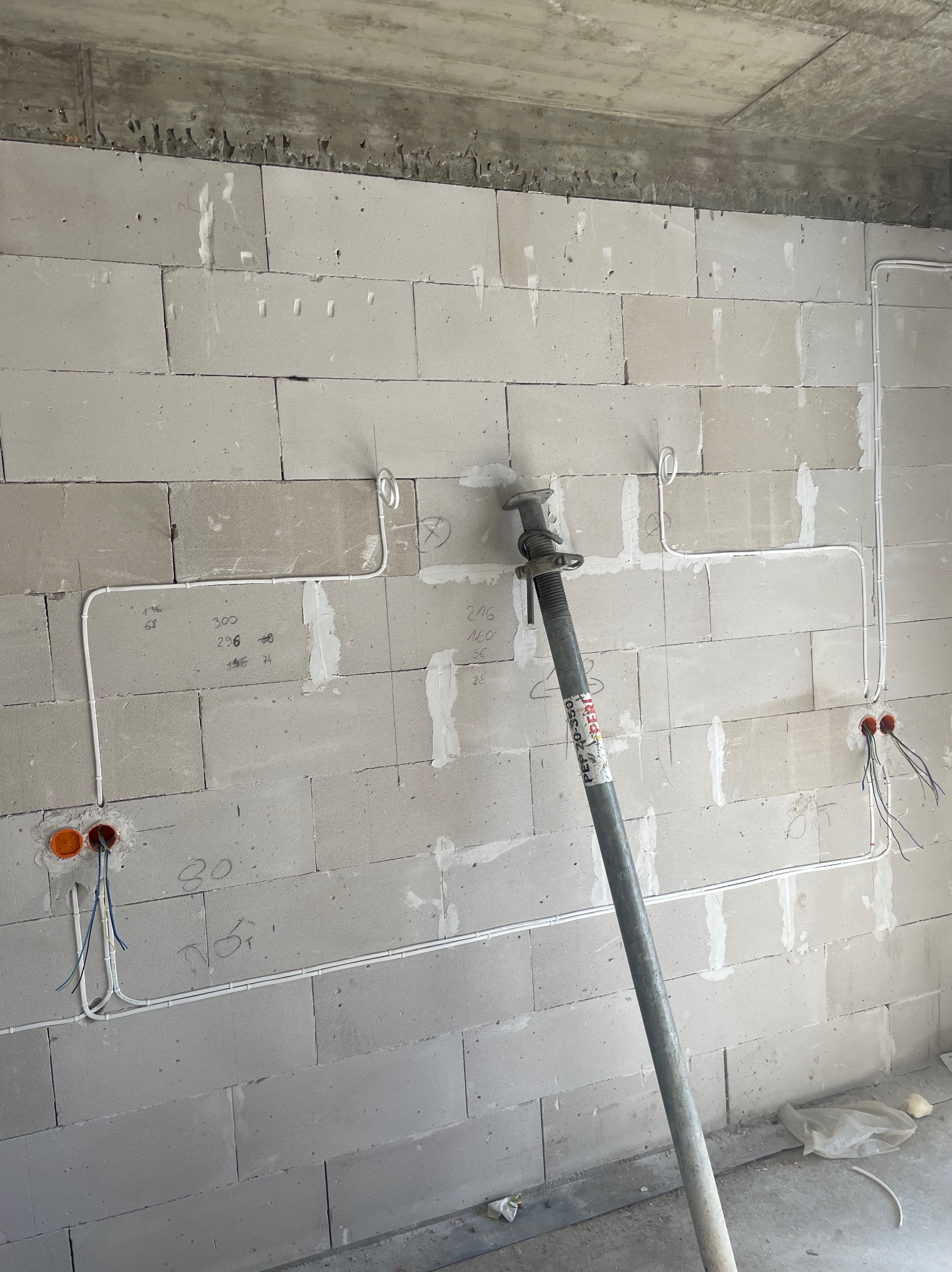 Ściana z betonu komórkowego w trakcie prac elektrycznych: widoczne kable w peszlach, puszki instalacyjne, podpory stropowe i odręczne notatki na ścianie.