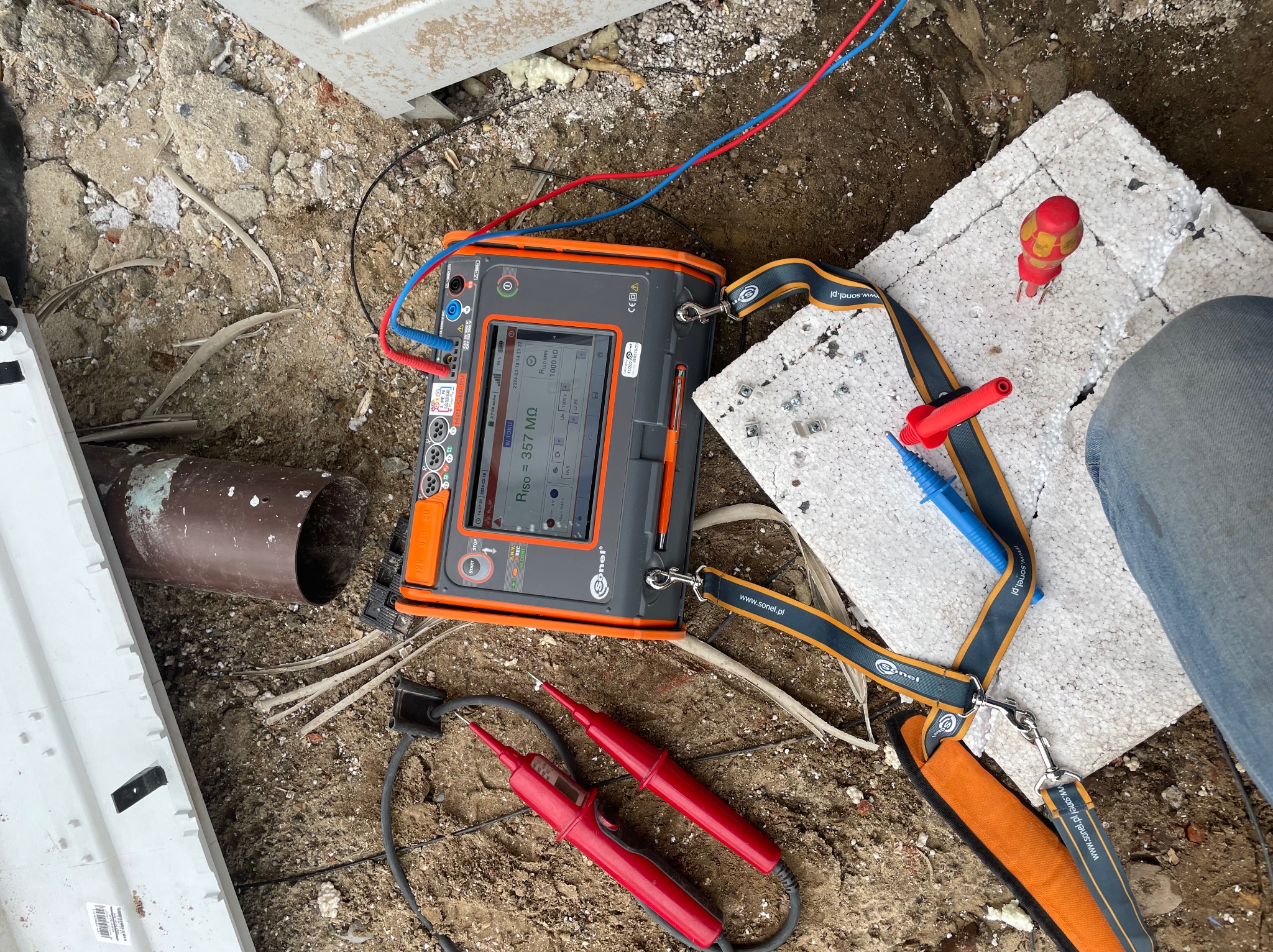 Miernik rezystancji izolacji Sonel na placu budowy, z przewodami pomiarowymi i sondami, wskazujący wynik pomiaru 357 MΩ, obok fragment styropianu i rury kanalizacyjnej.