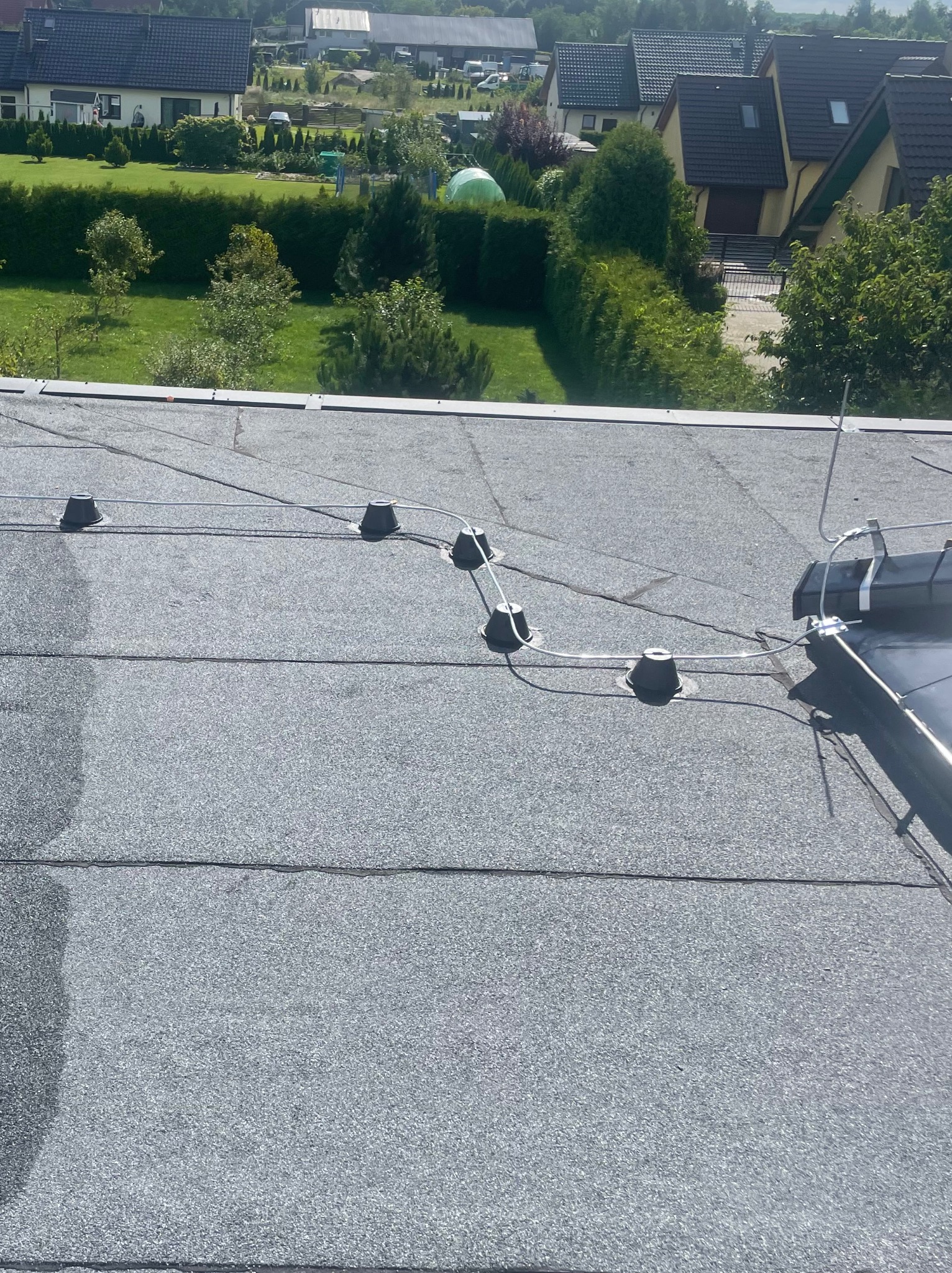 Instalacja odgromowa na dachu pokrytym papą, widok z góry na elementy mocujące i przewody, w tle zabudowa jednorodzinna i zieleń.