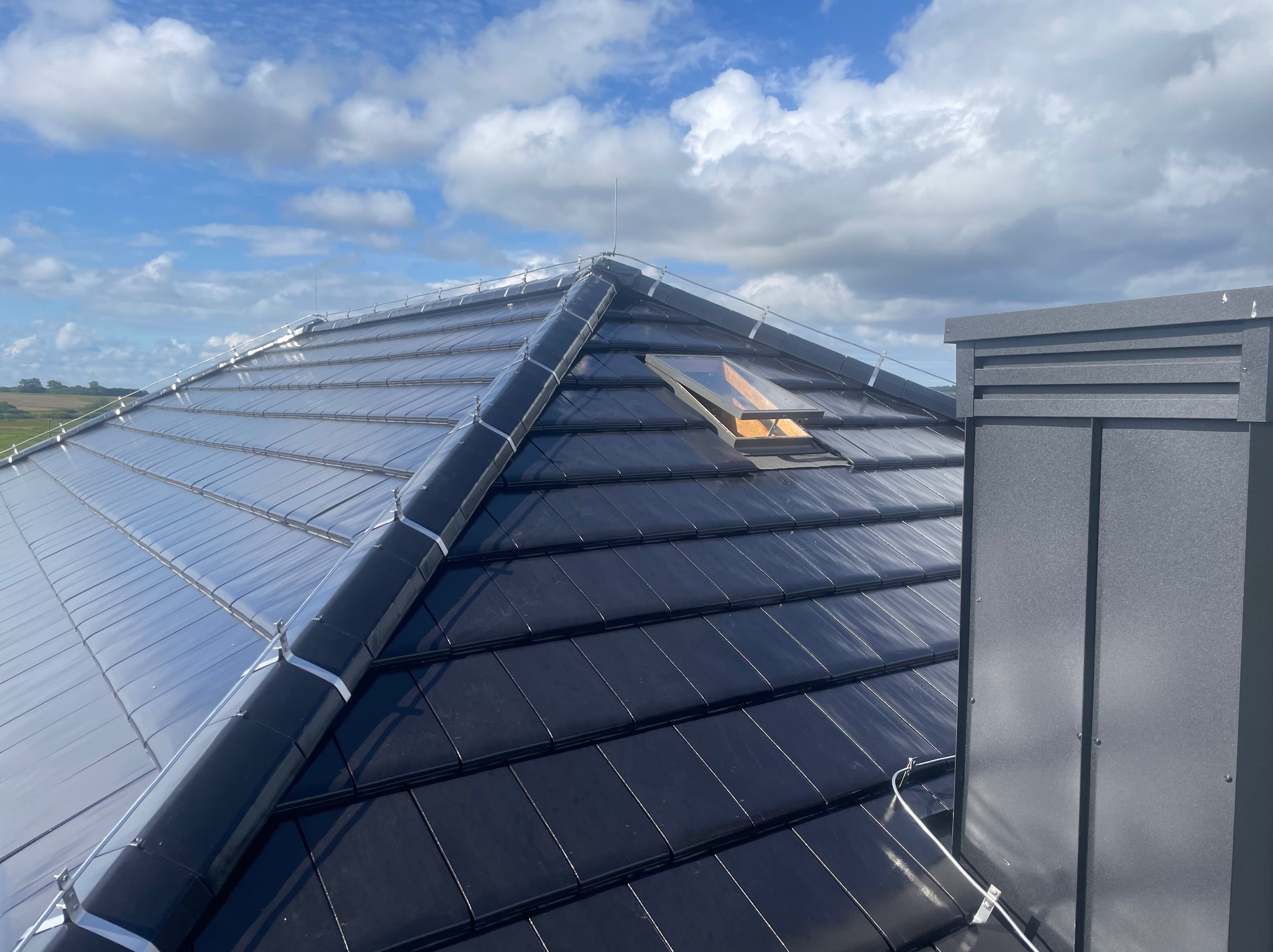 Czarny dach z dachówki ceramicznej z oknem połaciowym i instalacją odgromową na tle błękitnego nieba z chmurami. Widoczny komin.