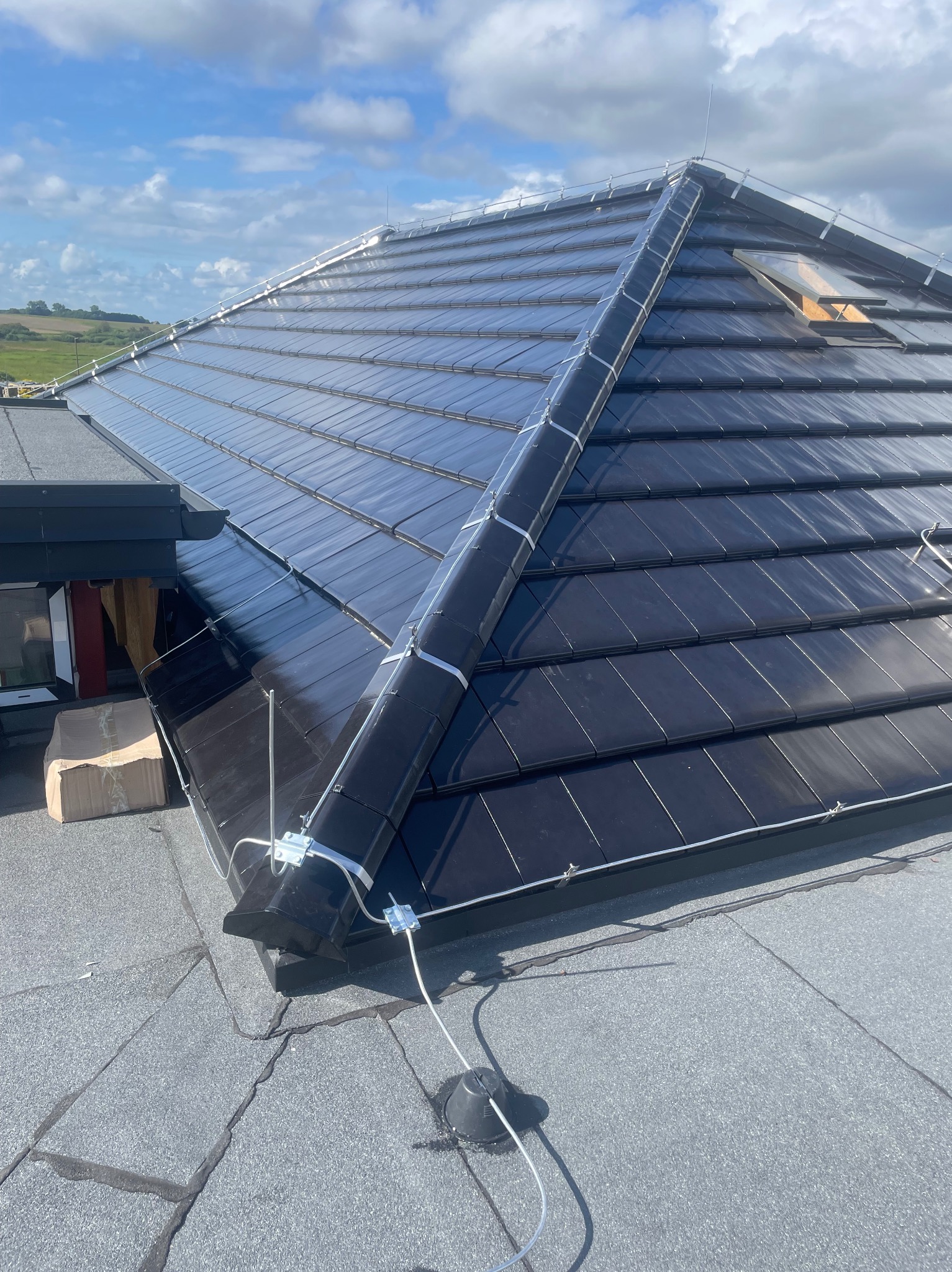 Czarny dach z dachówki ceramicznej z zamontowaną instalacją odgromową, zabezpieczoną białymi uchwytami. Widoczne przewody i elementy instalacji. W tle zielone wzgórza.