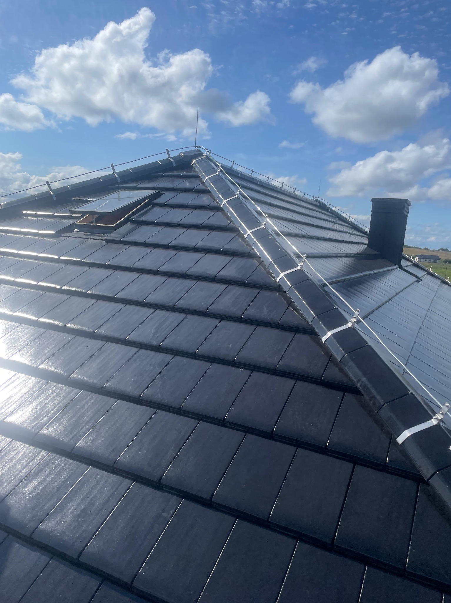 Czarny dach z dachówki ceramicznej, widoczne okno dachowe, komin i instalacja odgromowa z metalowymi uchwytami. Błękitne niebo z chmurami w tle.