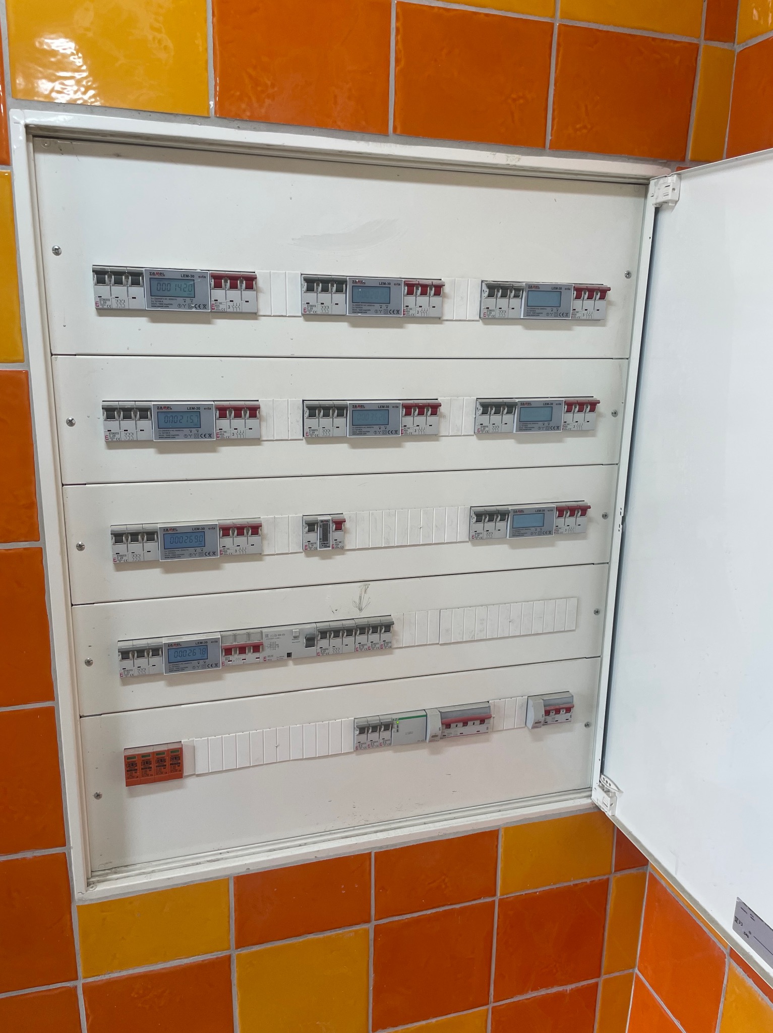 Rozdzielnia elektryczna z licznikami, zamontowana na ścianie wyłożonej pomarańczowo-żółtymi płytkami. Drzwiczki szafki otwarte, widoczne zabezpieczenia i elementy instalacji.