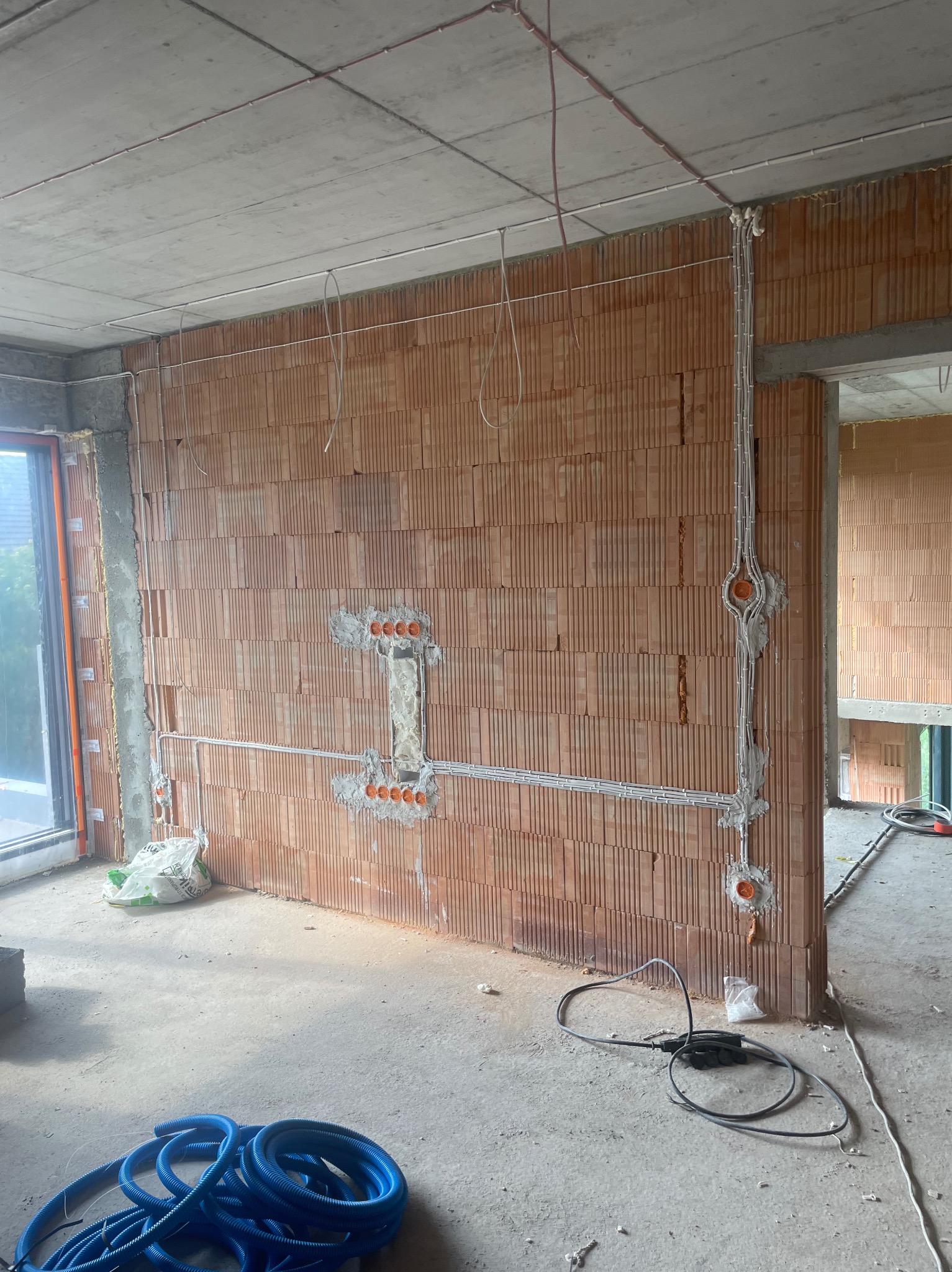 Instalacja elektryczna w surowym budynku: przewody w peszlach, puszki podtynkowe w ścianie z cegły dziurawki, widoczne kable na suficie i podłodze. Etap prac.