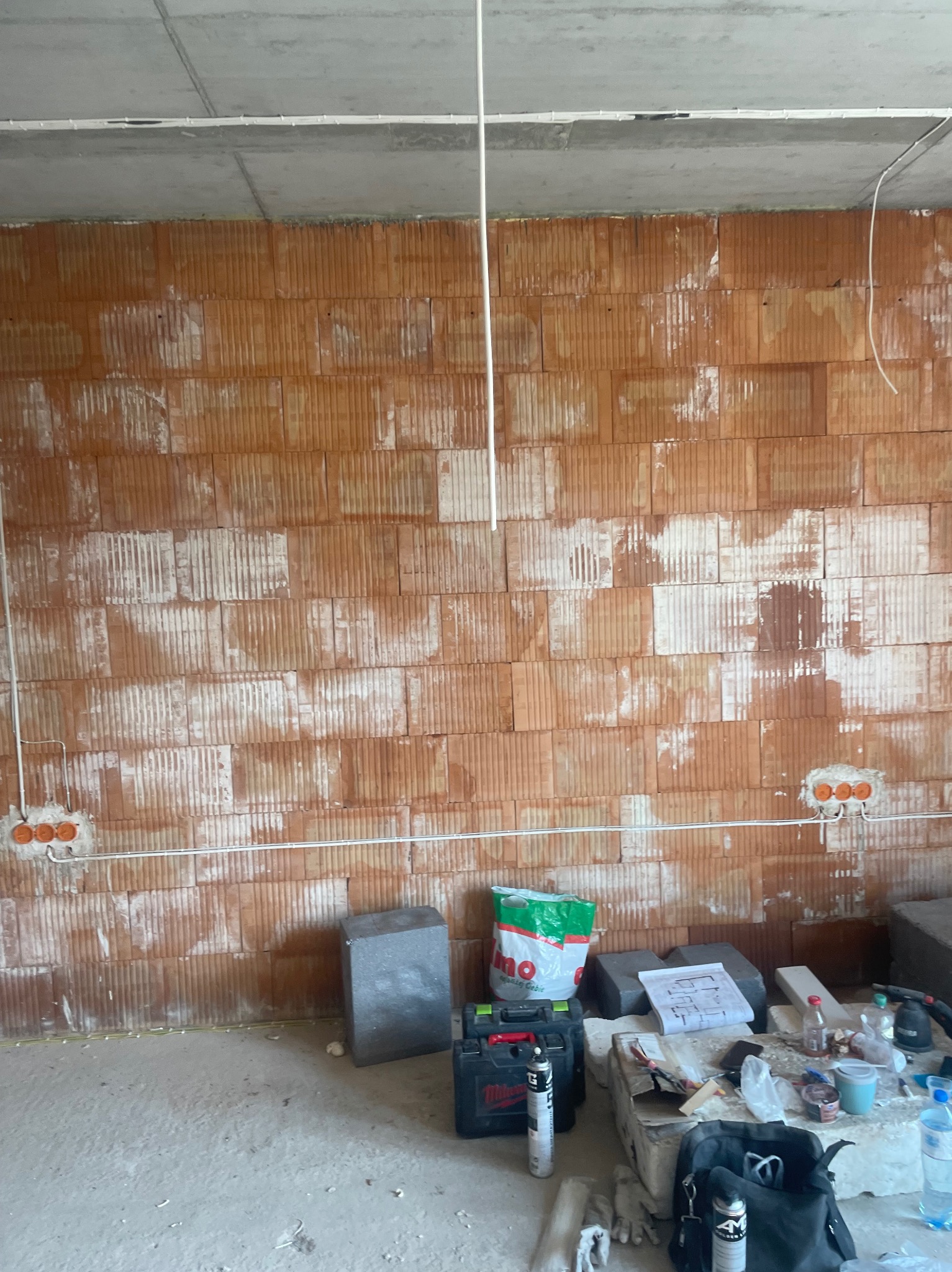 Surowa ściana z cegły z puszkami instalacyjnymi i przewodami. Widoczne narzędzia i materiały budowlane. Stan surowy przed tynkowaniem. Prace elektryczne w trakcie.