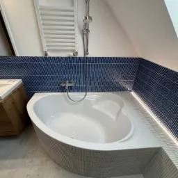 Nowoczesna łazienka na poddaszu z wanną narożną obudowaną mozaiką, niebieskimi płytkami na ścianach i grzejnikiem drabinkowym. Oświetlenie LED podkreśla krawędzie.