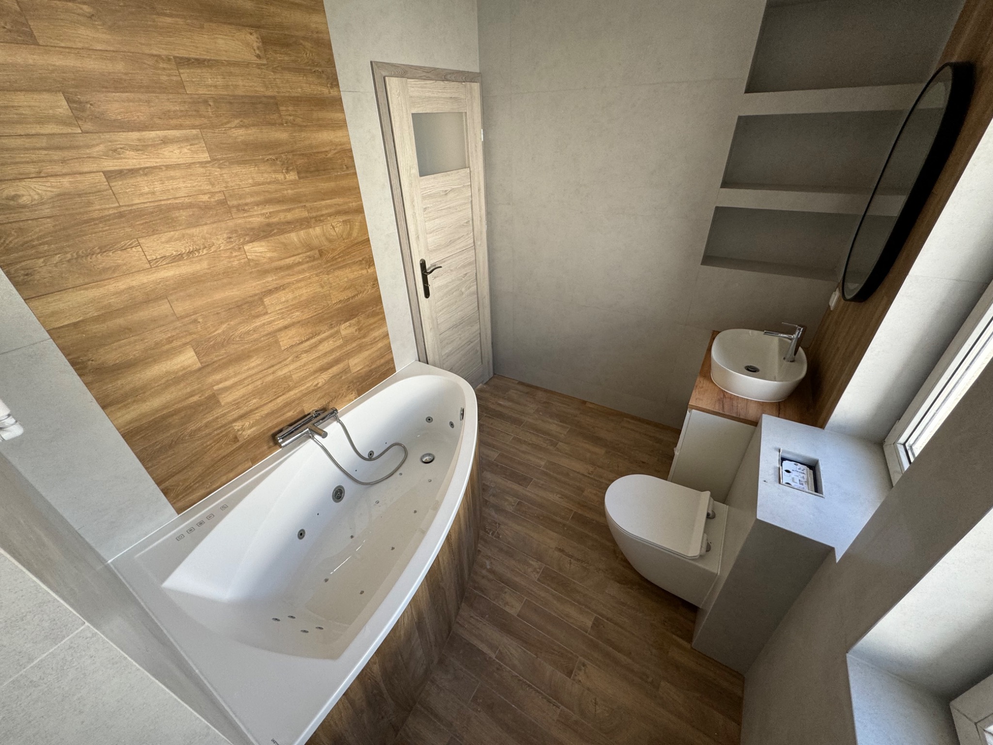 Nowoczesna łazienka z drewnopodobną podłogą i ścianą, biała wanna z hydromasażem, podwieszana toaleta i umywalka na drewnianym blacie, oświetlenie punktowe i minimalistyczne lustro.