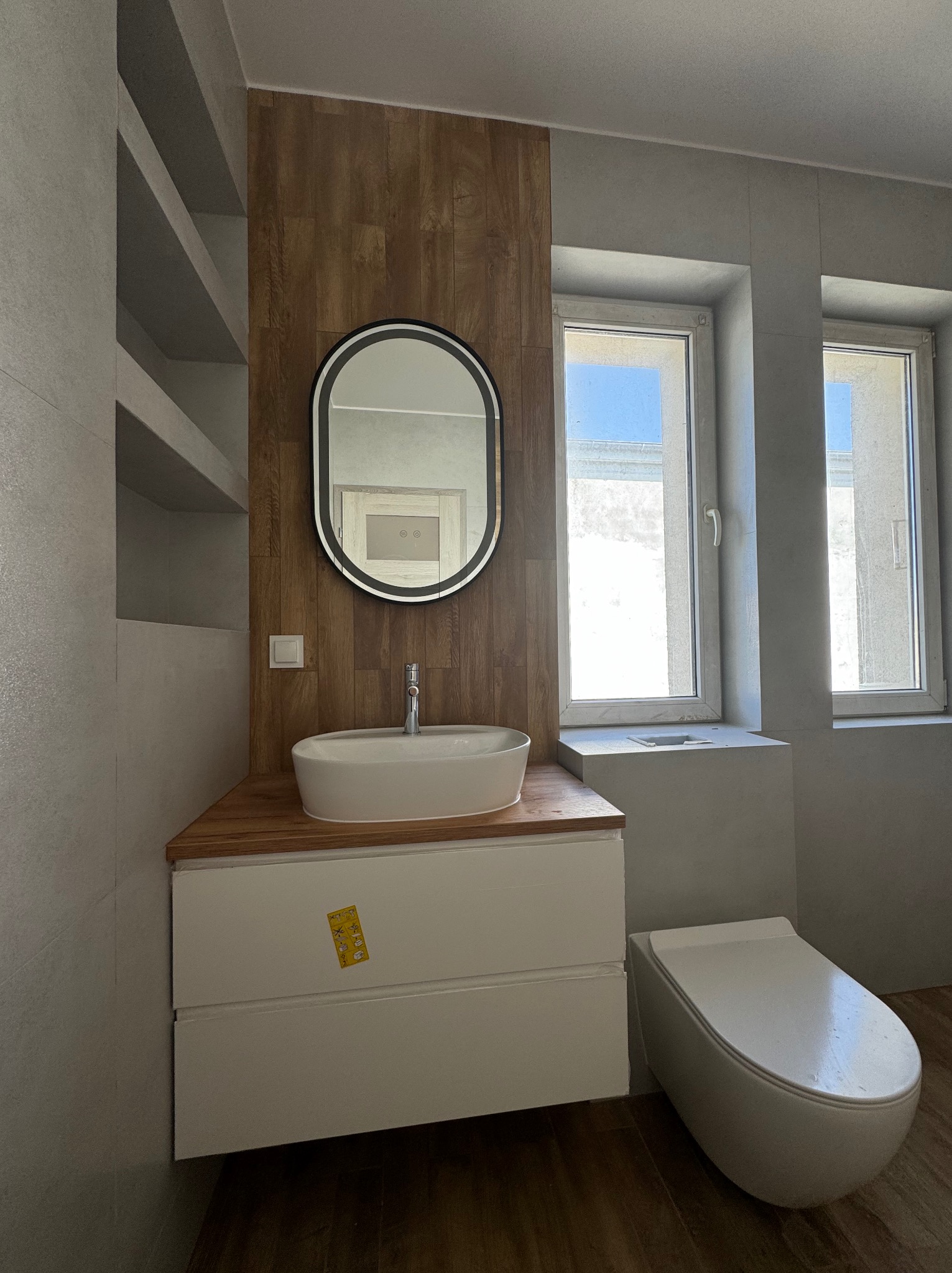 Nowoczesna łazienka z owalnym lustrem, drewnianą ścianą akcentującą i białą szafką pod umywalką z naklejkami, widok na toaletę i dwa okna.