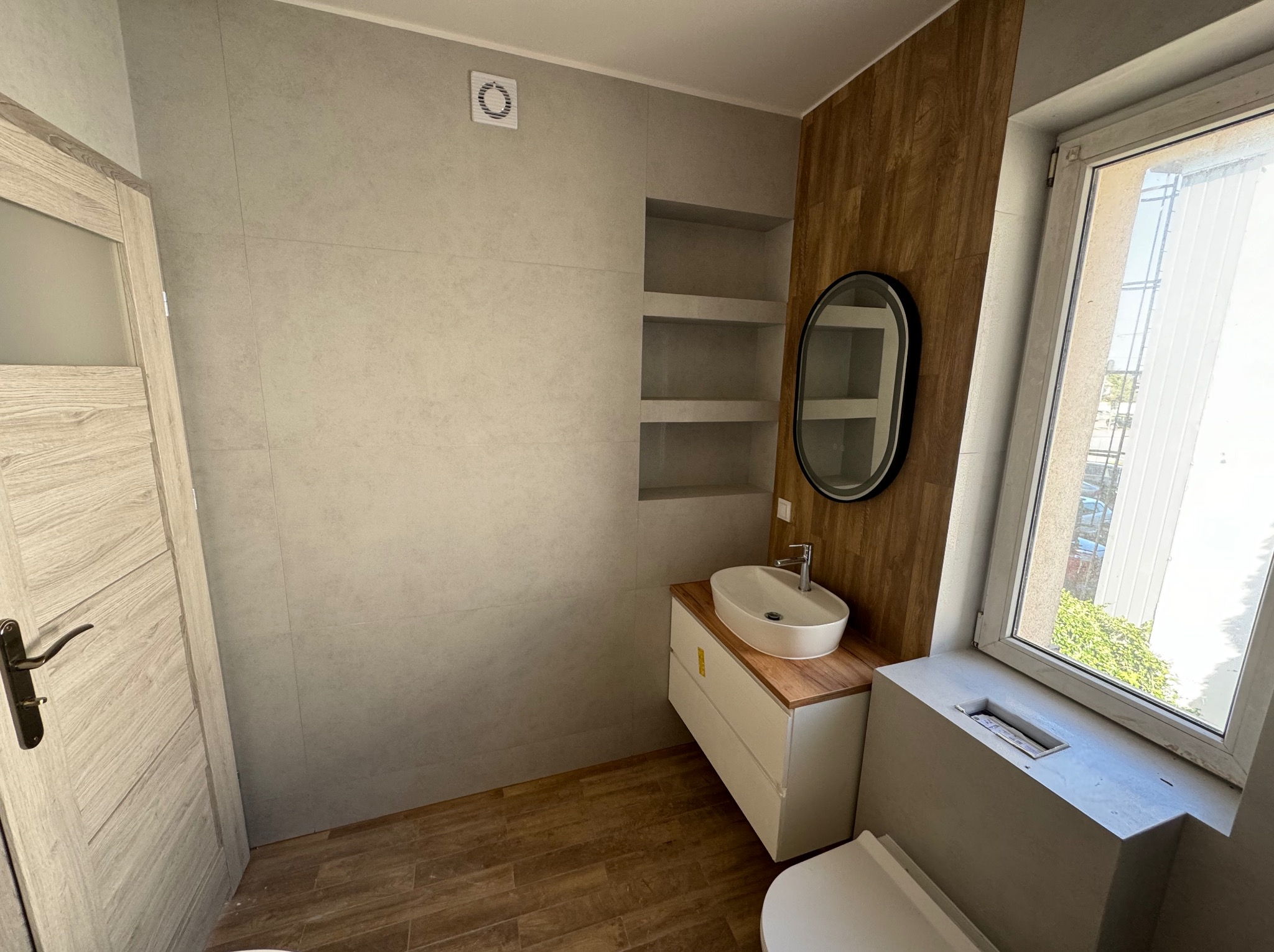 Jasna łazienka z szarymi płytkami, drewnopodobną ścianą, owalną umywalką na szafce z drewnianym blatem, wbudowanymi półkami i drewnianą podłogą.