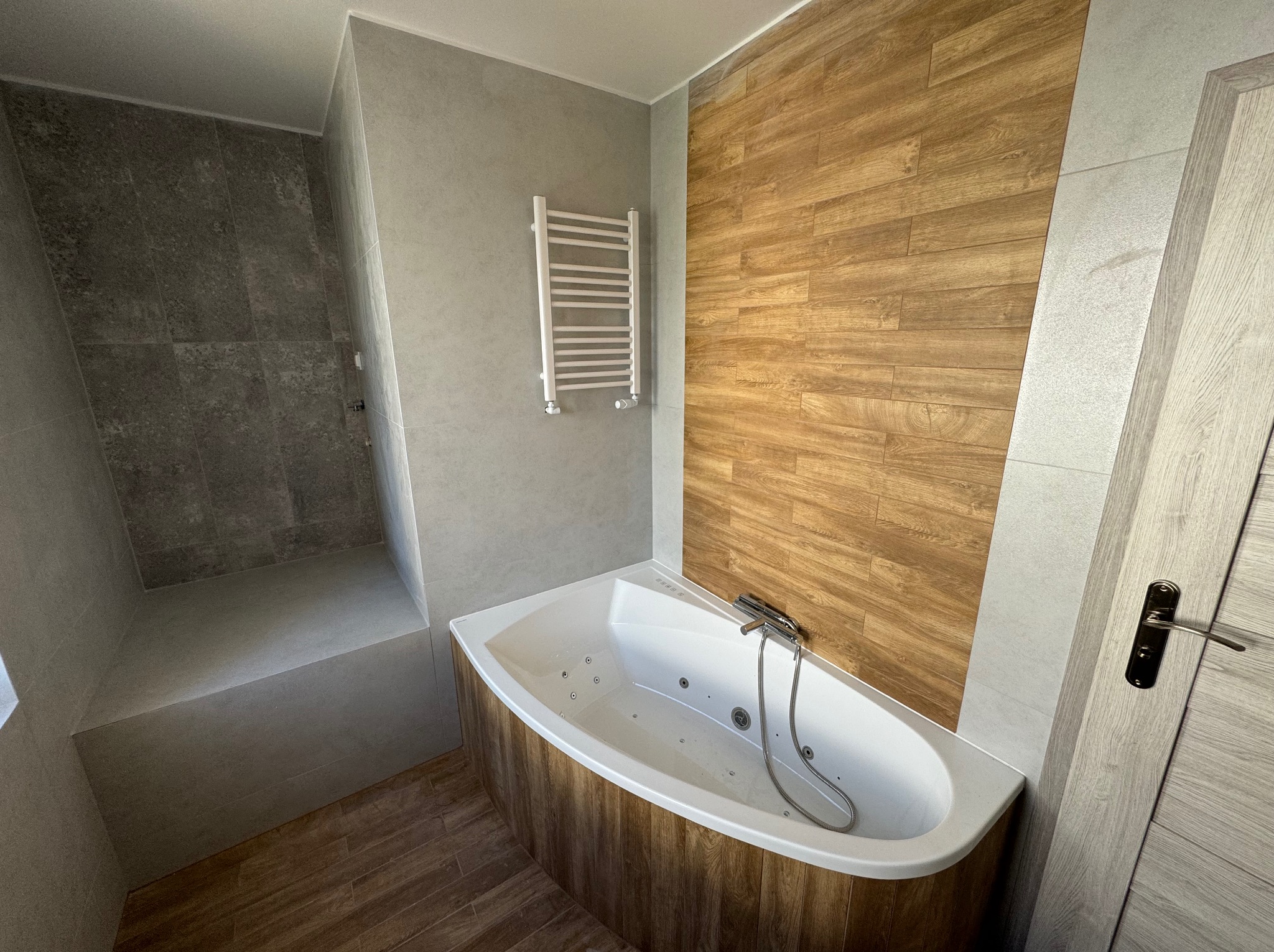 Nowoczesna łazienka z narożną wanną z hydromasażem obudowaną panelami drewnopodobnymi, szarymi płytkami na ścianach i podłodze, grzejnikiem drabinkowym i kabiną prysznicową walk-in.