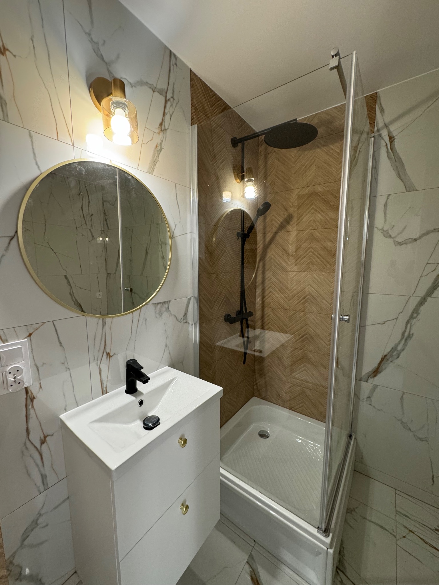 Nowoczesna łazienka z kabiną prysznicową, umywalką z szafką, okrągłym lustrem w złotej ramie i marmurowymi płytkami na ścianach.