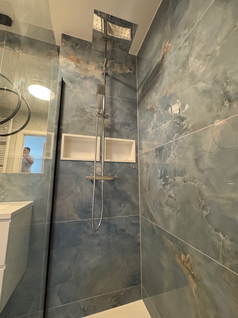 Wykończona łazienka z niebieskimi płytkami imitującymi marmur, chromowana armatura prysznicowa, wnęki ścienne i odbicie osoby w lustrze.