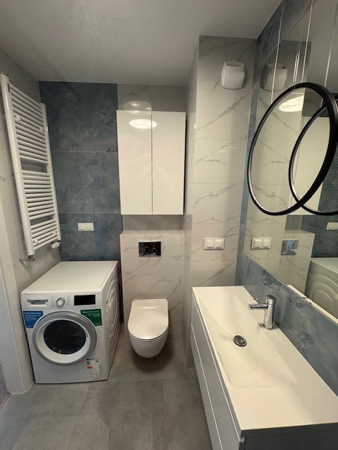 Nowoczesna łazienka z szarymi płytkami imitującymi marmur, białą zabudową umywalki z prostokątną umywalką, podwieszaną toaletą, pralką i grzejnikiem drabinkowym.