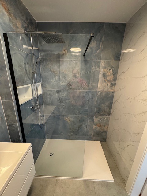 Nowoczesna kabina prysznicowa typu walk-in z odpływem liniowym, wykończona płytkami imitującymi marmur w odcieniach szarości i błękitu, z widocznym prysznicem deszczownicą oraz wnęką na kosmetyki.