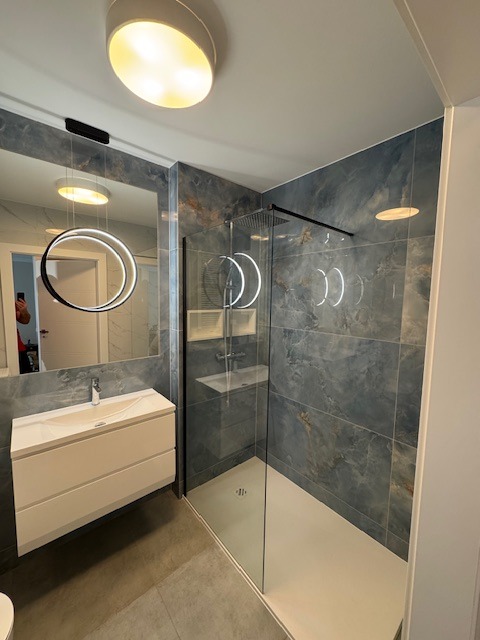 Nowoczesna łazienka z kabiną prysznicową typu walk-in, umywalką nablatową na szafce i lustrem z podświetleniem LED, wykończona płytkami imitującymi marmur w odcieniach szarości i błękitu.