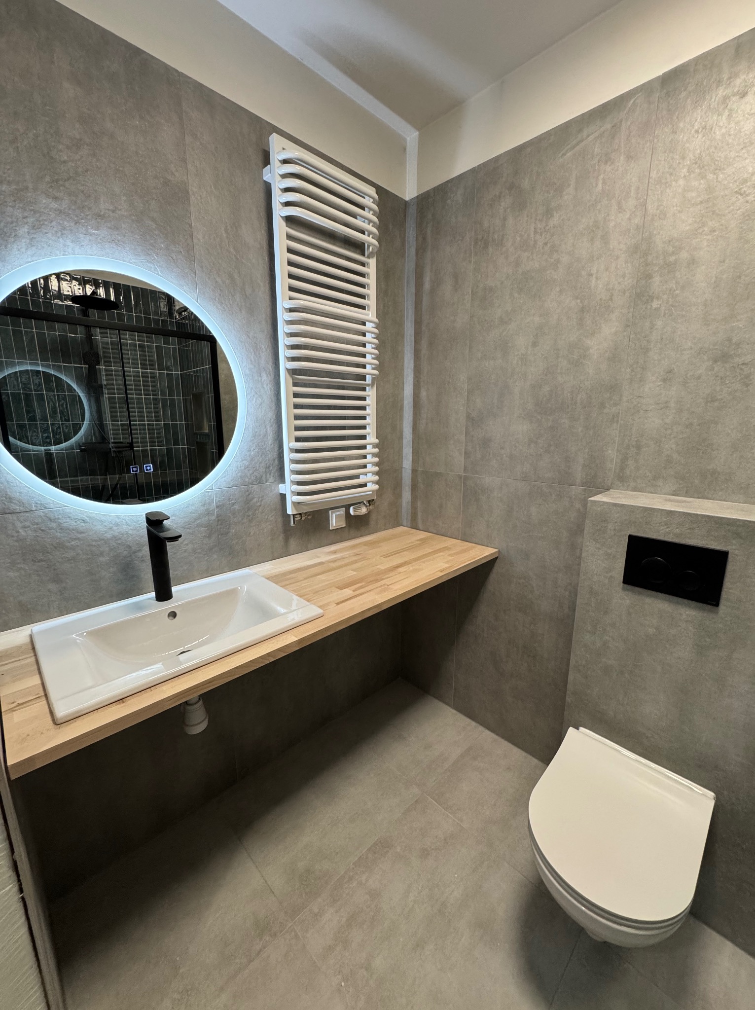 Nowoczesna łazienka w szarościach z drewnianym blatem, okrągłe lustro z podświetleniem LED, biała umywalka z czarną baterią, grzejnik drabinkowy i podwieszana toaleta.