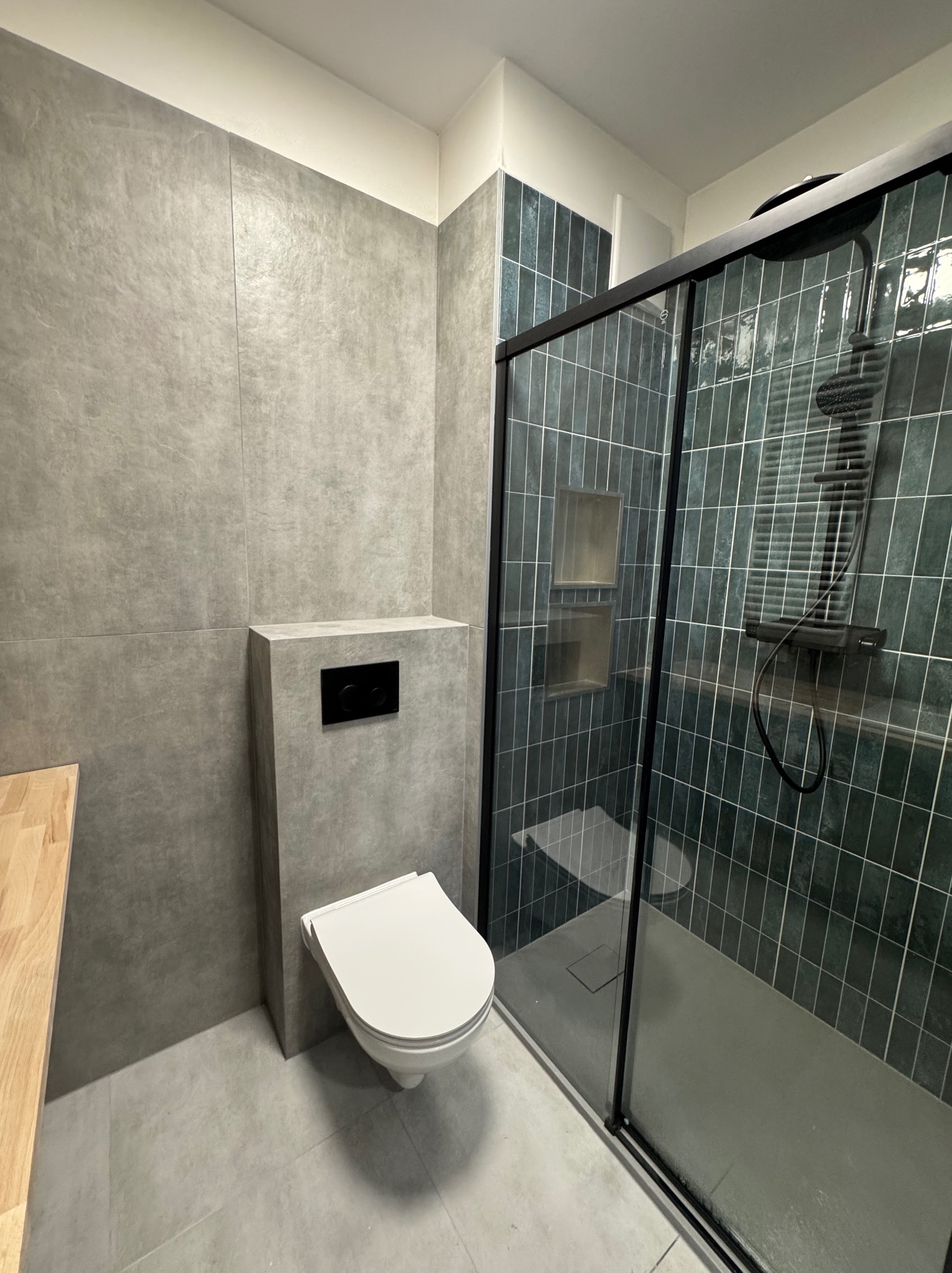 Nowoczesna łazienka z szarymi płytkami imitującymi beton, wisząca toaleta z czarnym panelem sterującym, kabina prysznicowa typu walk-in z ciemnozielonymi płytkami w pionowy wzór i wnękami...