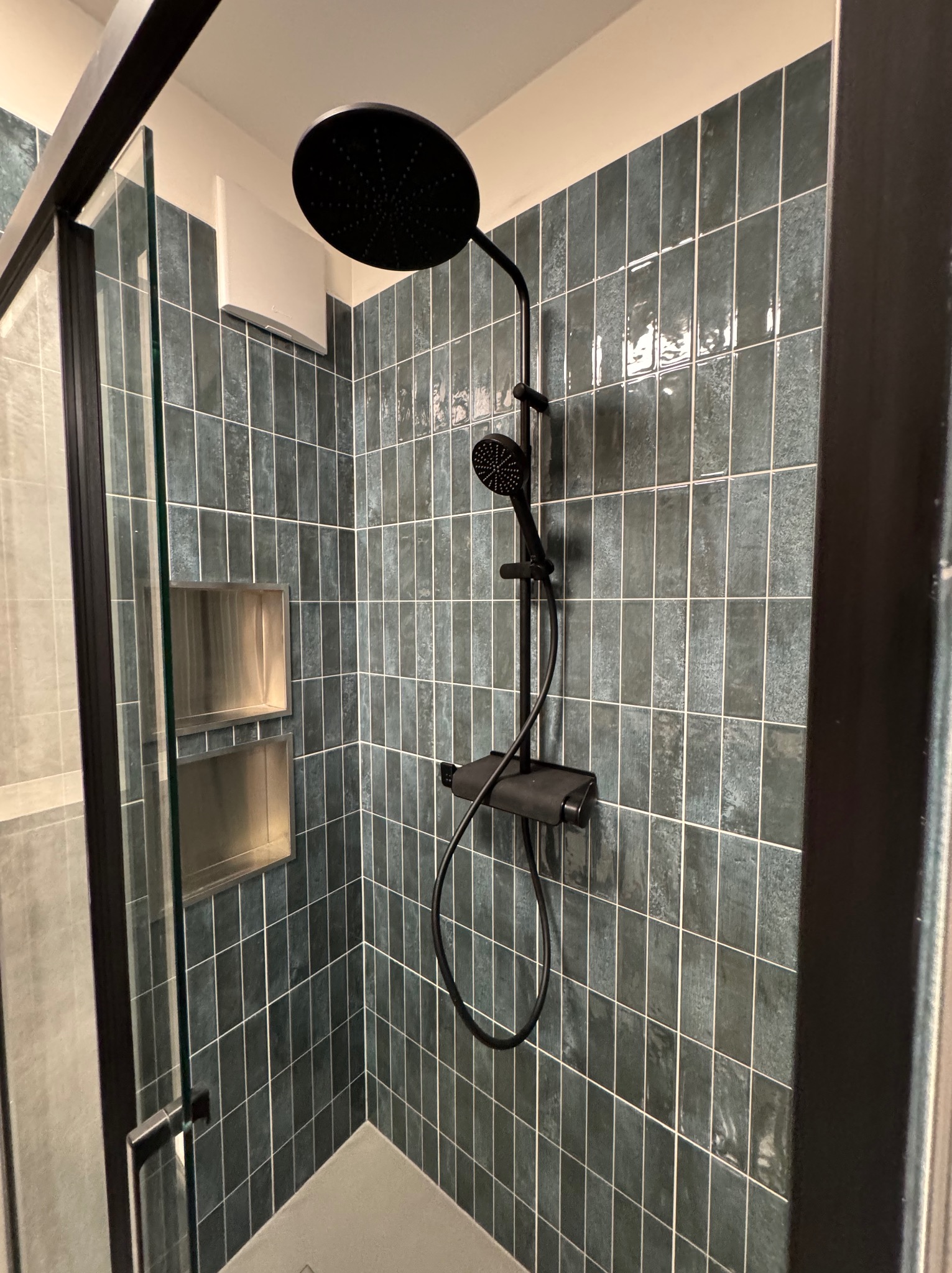 Nowoczesna kabina prysznicowa z czarną armaturą, wyłożona pionowymi, błyszczącymi, zielono-niebieskimi płytkami o nieregularnej fakturze, z wnękami w ścianie wykończonymi metalem.