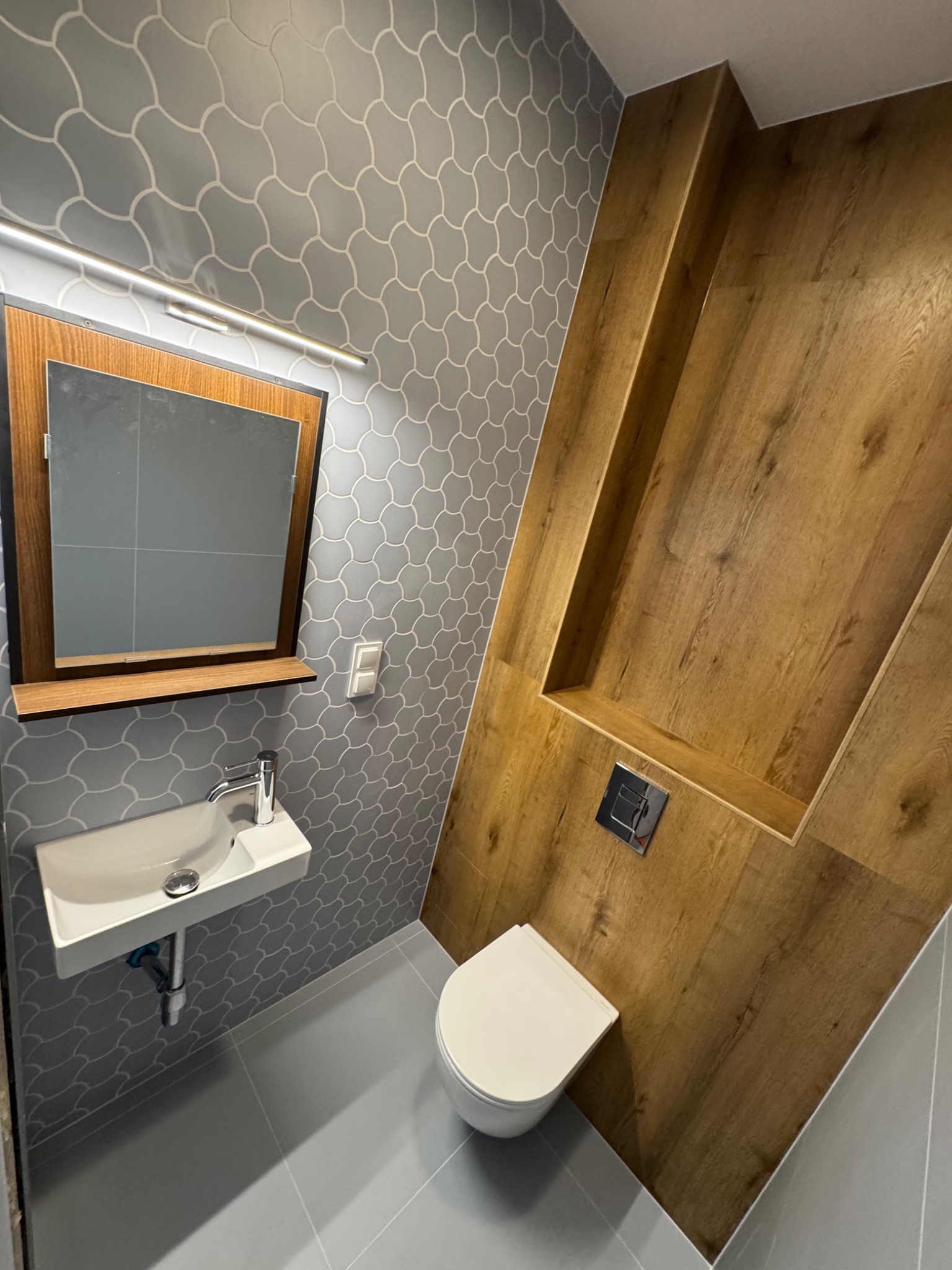 Nowoczesna łazienka z szarymi płytkami o wzorze łuski, drewnianą ścianą z wnęką i białym, podwieszanym WC oraz umywalką z lustrem.