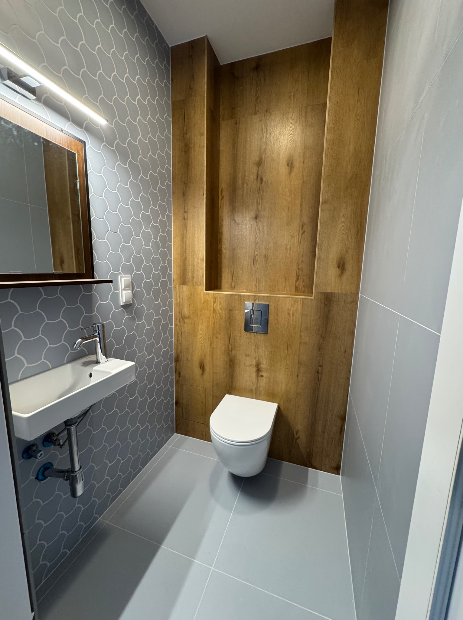 Nowoczesna łazienka z mozaiką w odcieniach szarości, drewnianą zabudową WC i minimalistyczną umywalką. Oświetlenie LED podkreśla detale.