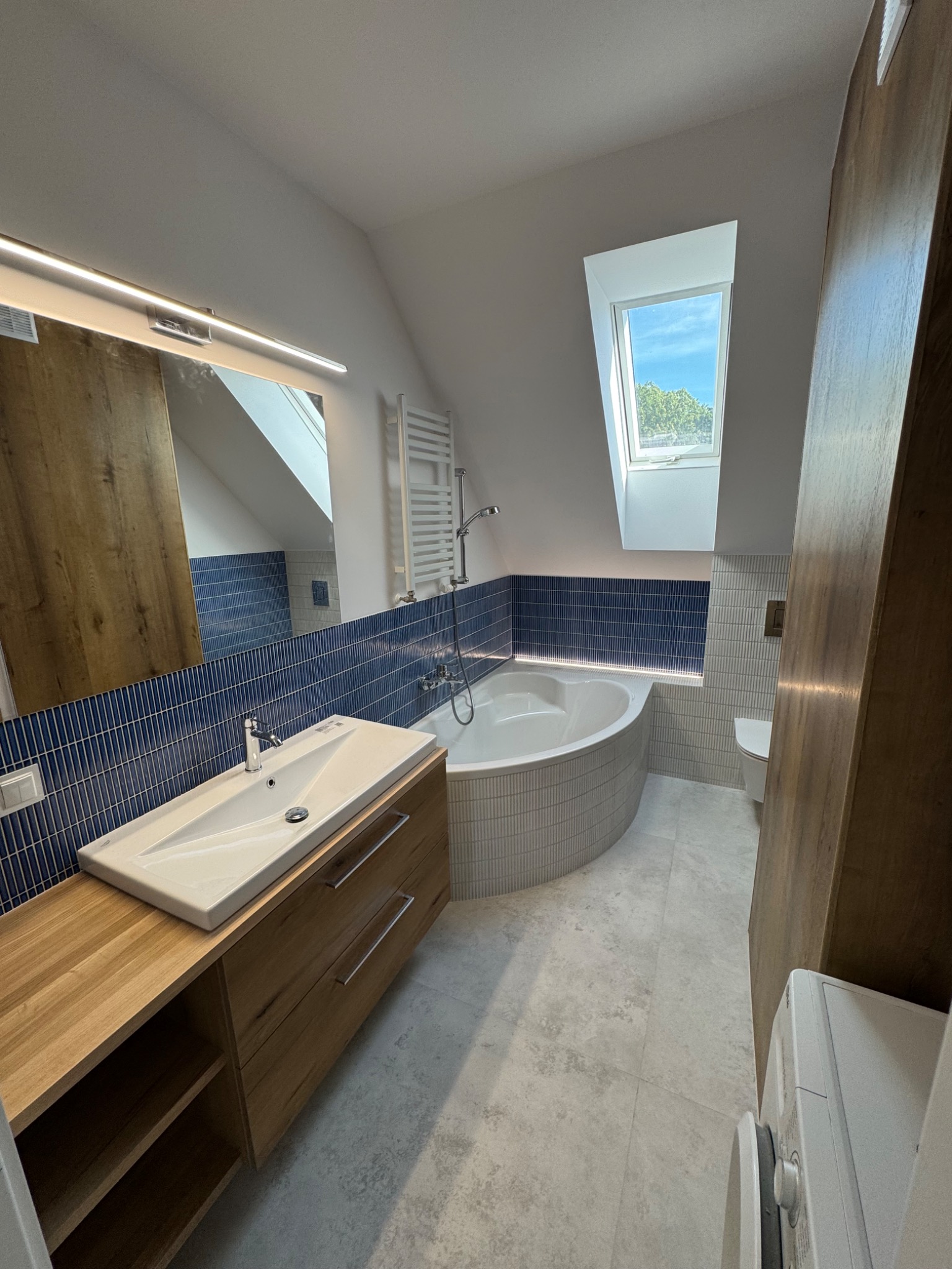 Stylowa łazienka na poddaszu z niebieską mozaiką, drewnianą szafką pod umywalką i owalną wanną z subtelnym oświetleniem LED. Widok na okno dachowe.