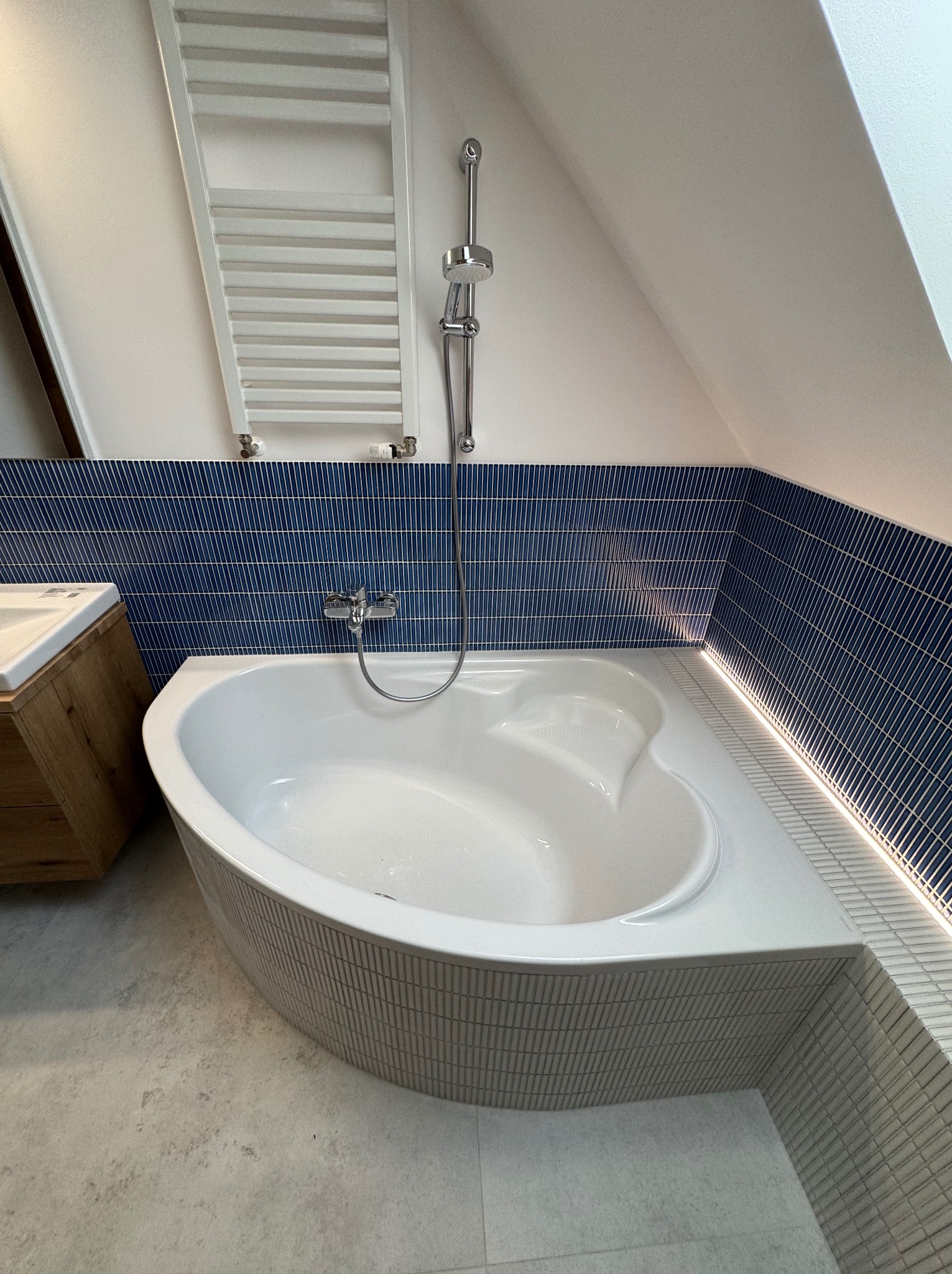 Nowoczesna łazienka na poddaszu z wanną narożną obudowaną mozaiką, niebieskimi płytkami na ścianach i grzejnikiem drabinkowym. Oświetlenie LED podkreśla krawędzie.