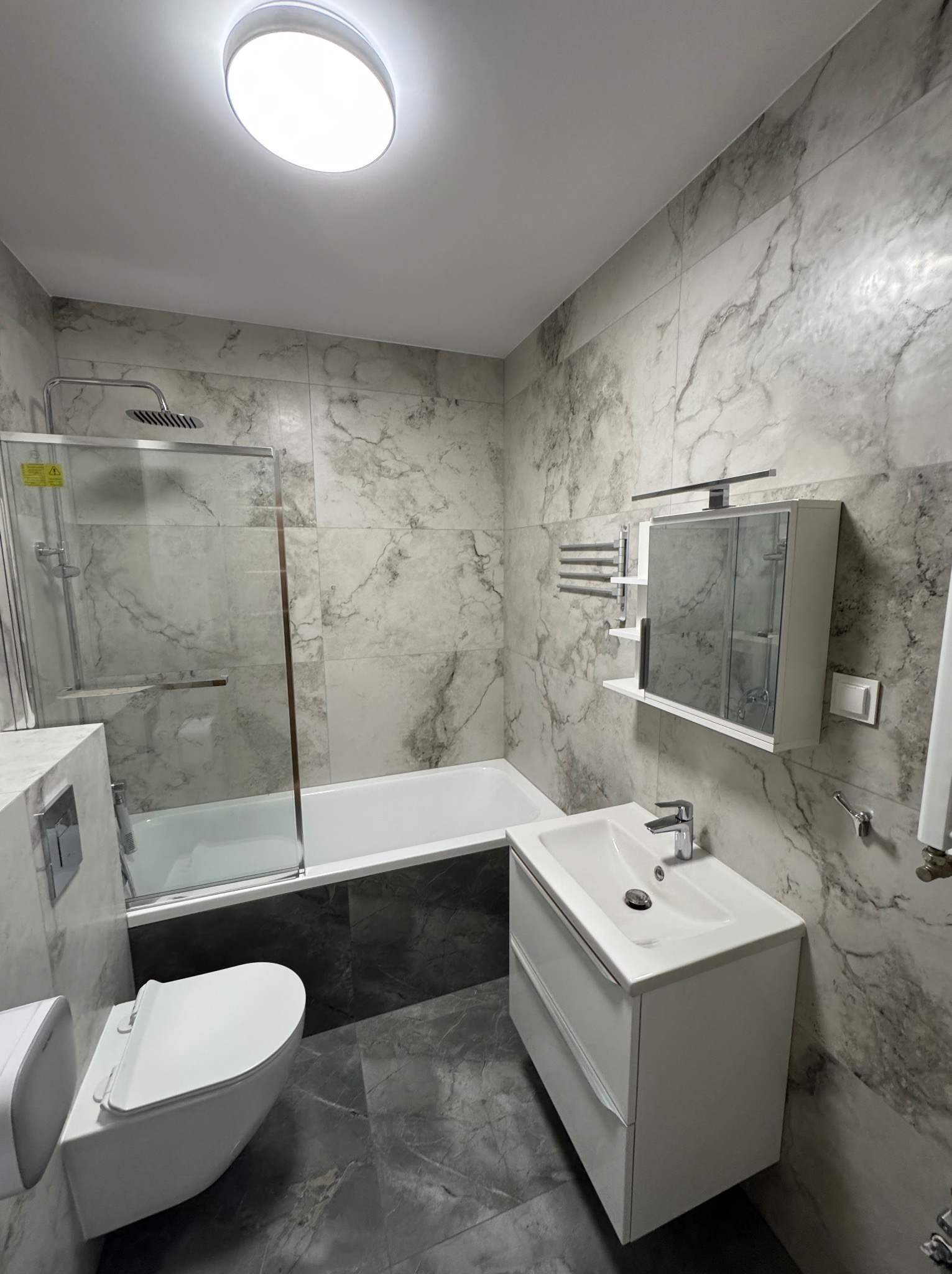 Nowoczesna łazienka z marmurowymi płytkami, białą armaturą i szklaną kabiną prysznicową. Umywalka z szafką, toaleta i grzejnik drabinkowy tworzą spójną całość.