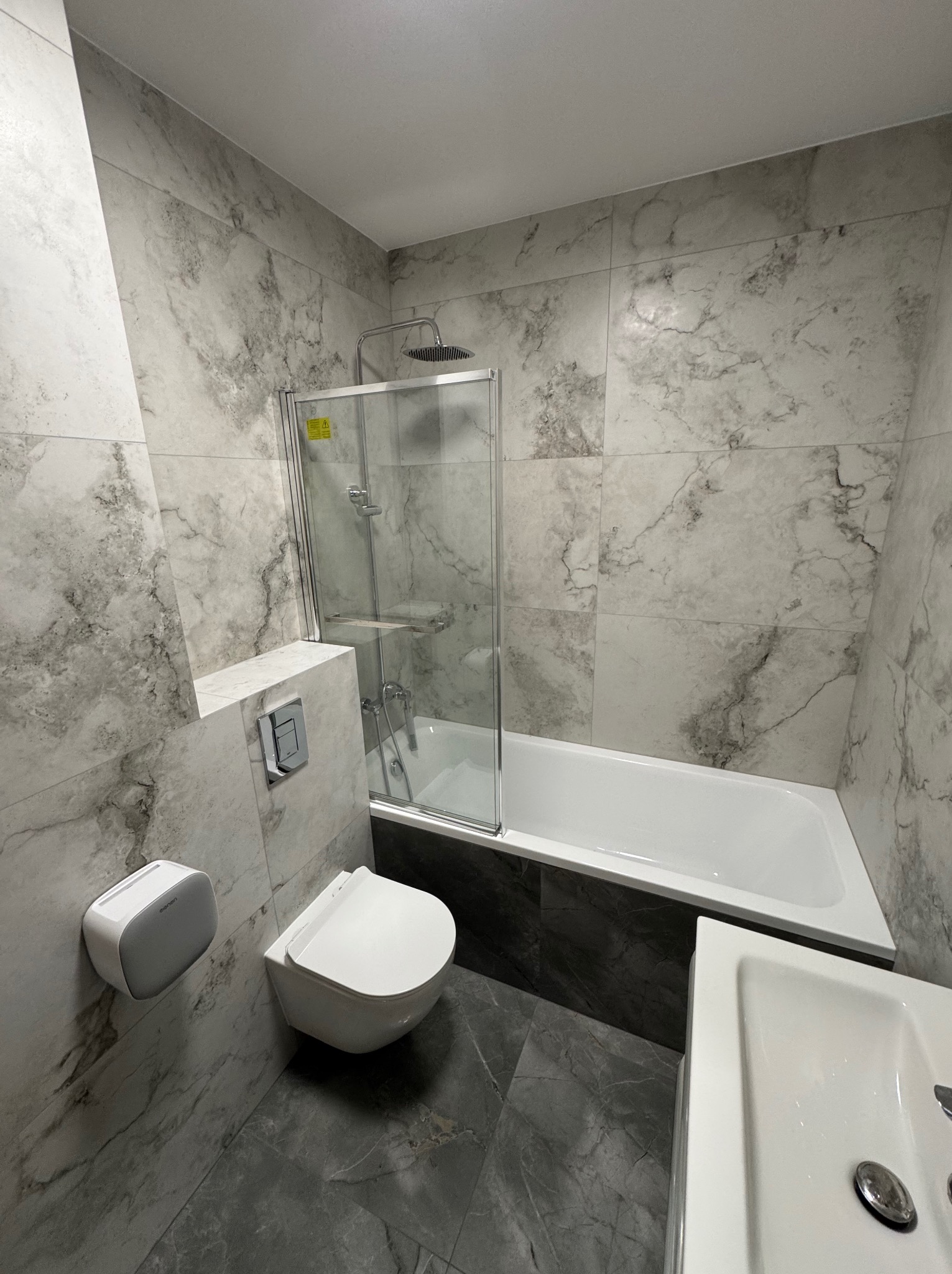 Nowoczesna łazienka z marmurowymi płytkami, kabiną prysznicową nad wanną, podwieszaną toaletą i umywalką. Szare płytki podłogowe. Minimalistyczny design.