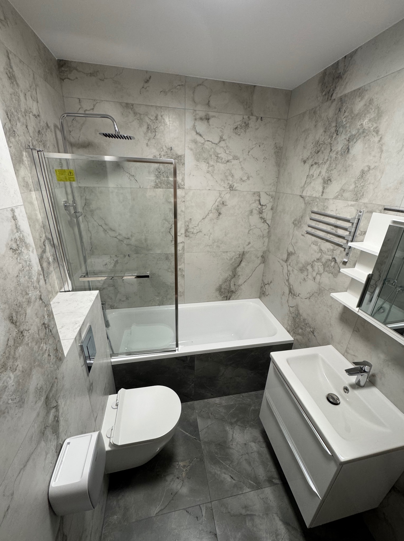 Nowoczesna łazienka z marmurowymi płytkami, kabiną prysznicową nad wanną, podwieszaną toaletą i umywalką z szafką. Minimalistyczny design, szaro-biała kolorystyka.