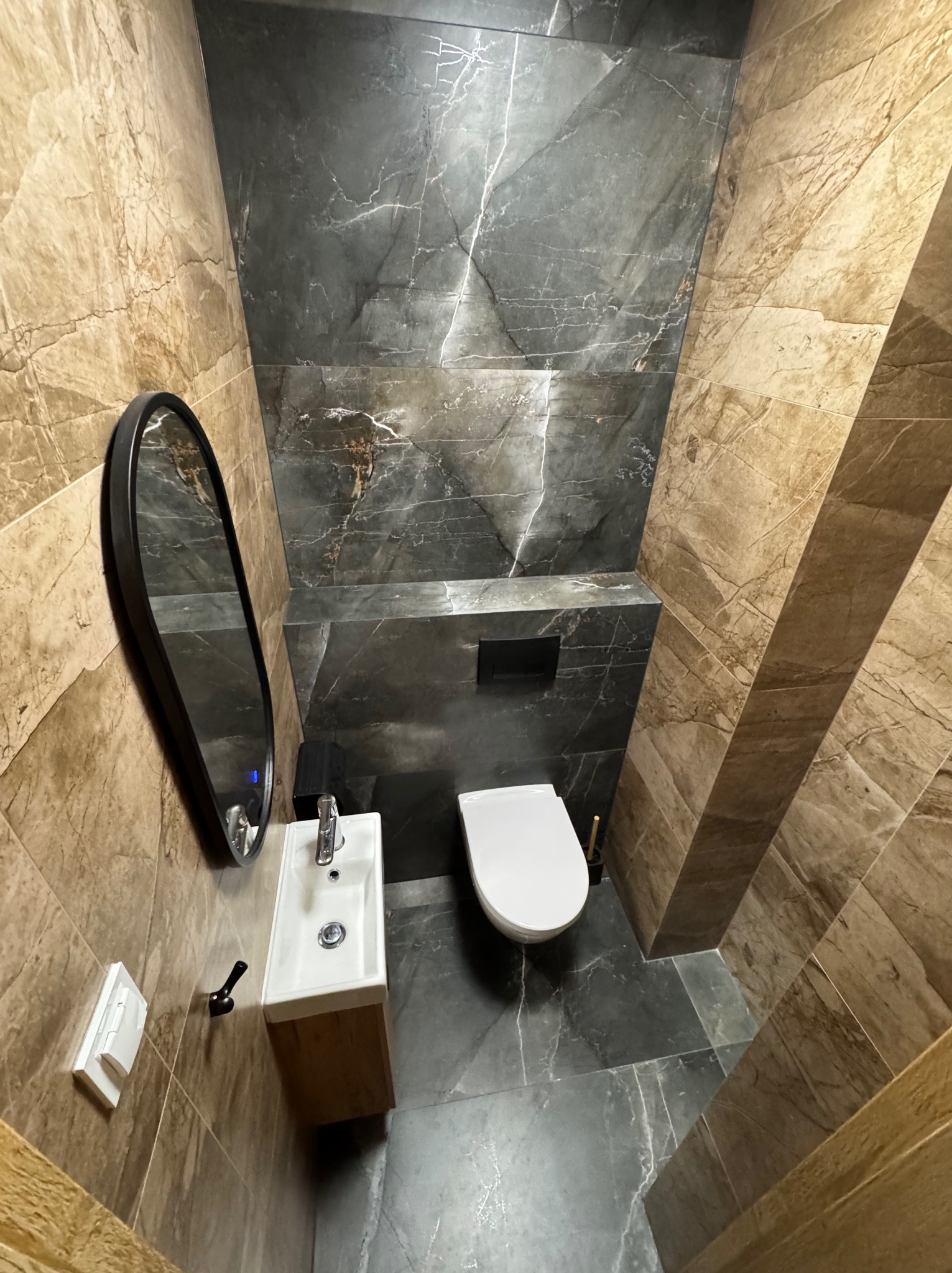 Nowoczesna łazienka z marmurowymi płytkami w odcieniach beżu i szarości. Umywalka z szafką, owalne lustro i toaleta tworzą spójną całość.