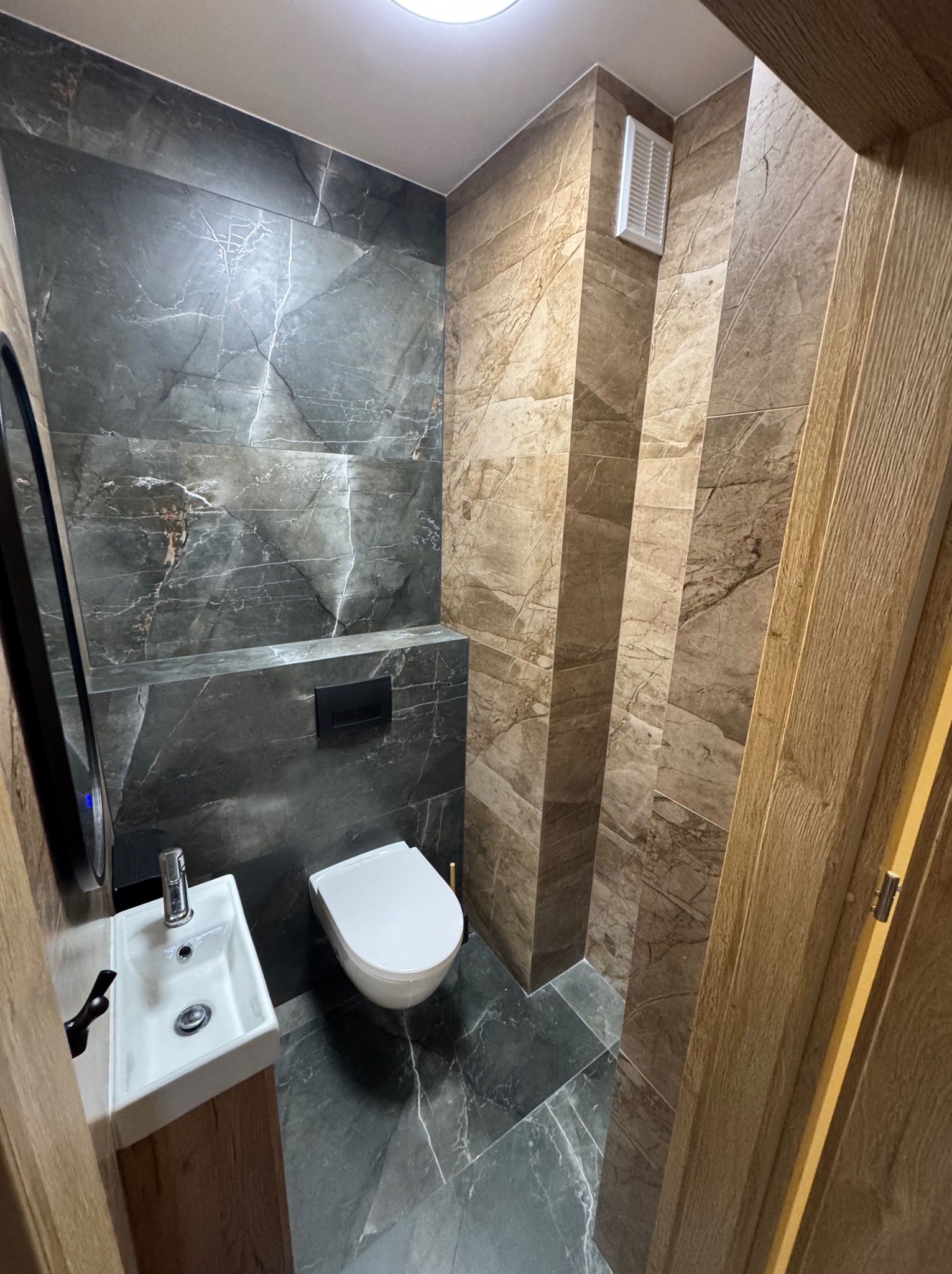Nowoczesna łazienka z marmurowymi płytkami w odcieniach szarości i brązu. Umywalka nablatowa, wisząca toaleta i drewniane drzwi tworzą spójną całość.