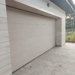 Zamknięta brama garażowa segmentowa w kolorze jasnego drewna, z poziomymi panelami, zamontowana w jasnym garażu z kostki brukowej na podjeździe.