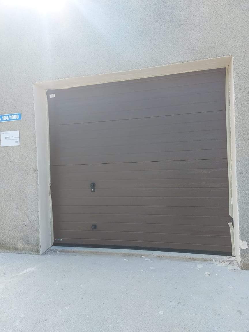 Zamknięta, brązowa brama garażowa segmentowa z dwoma uchwytami, osadzona w surowej, betonowej wnęce. Widoczna tabliczka adresowa po lewej stronie.
