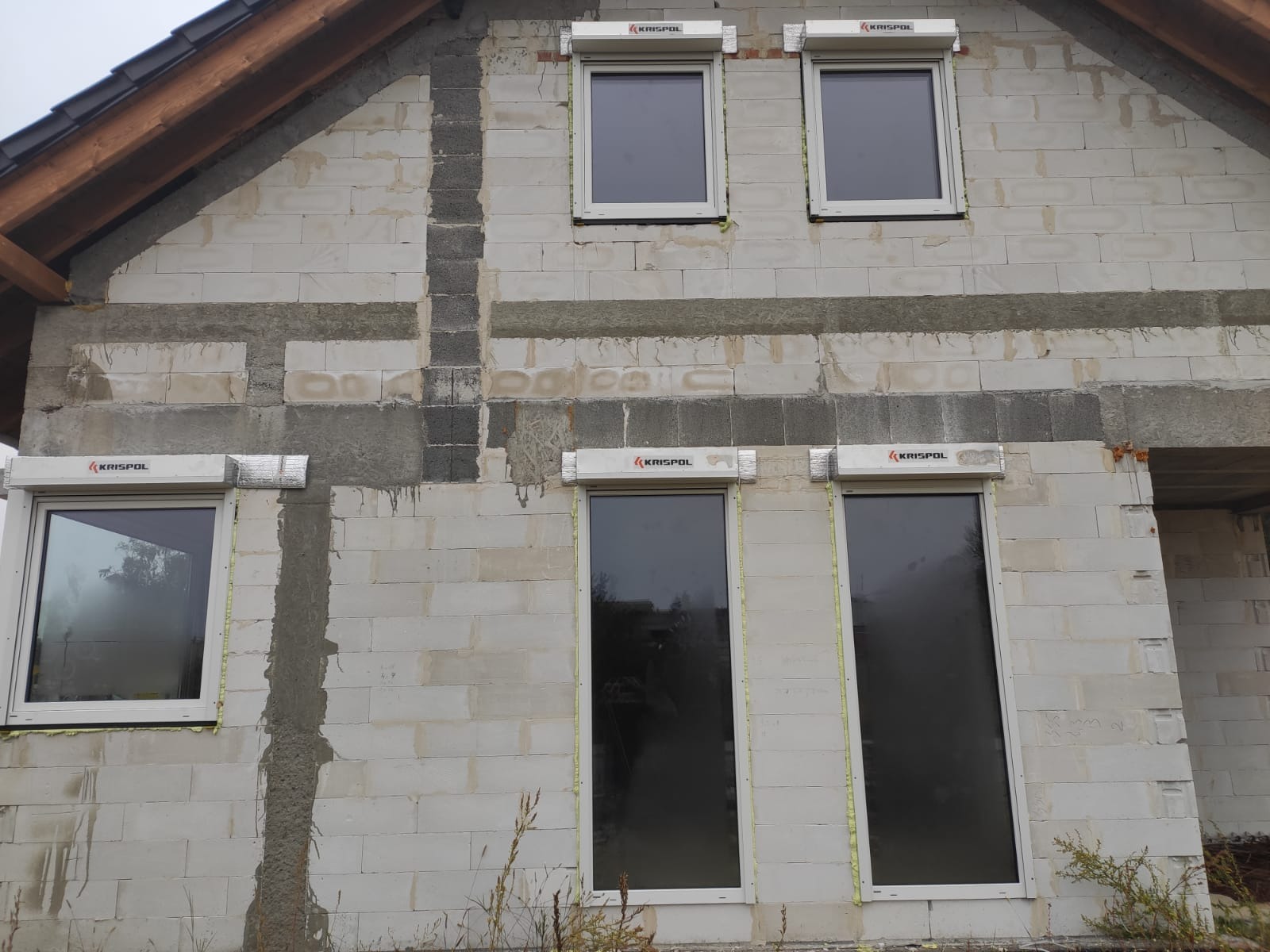 Surowy stan budynku z zamontowanymi oknami PCV i roletami zewnętrznymi marki Krispol, widoczne bloczki betonowe i ślady zaprawy.