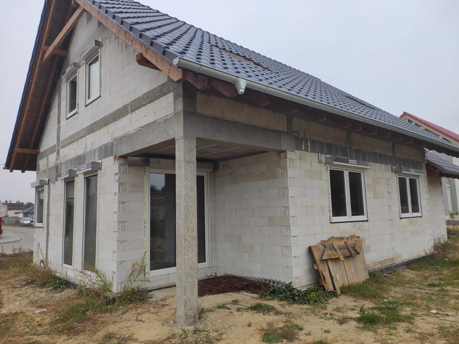 Niedokończony dom z białych bloczków, z zamontowanymi oknami i szarą dachówką, widoczne elementy konstrukcyjne pod rolety zewnętrzne.