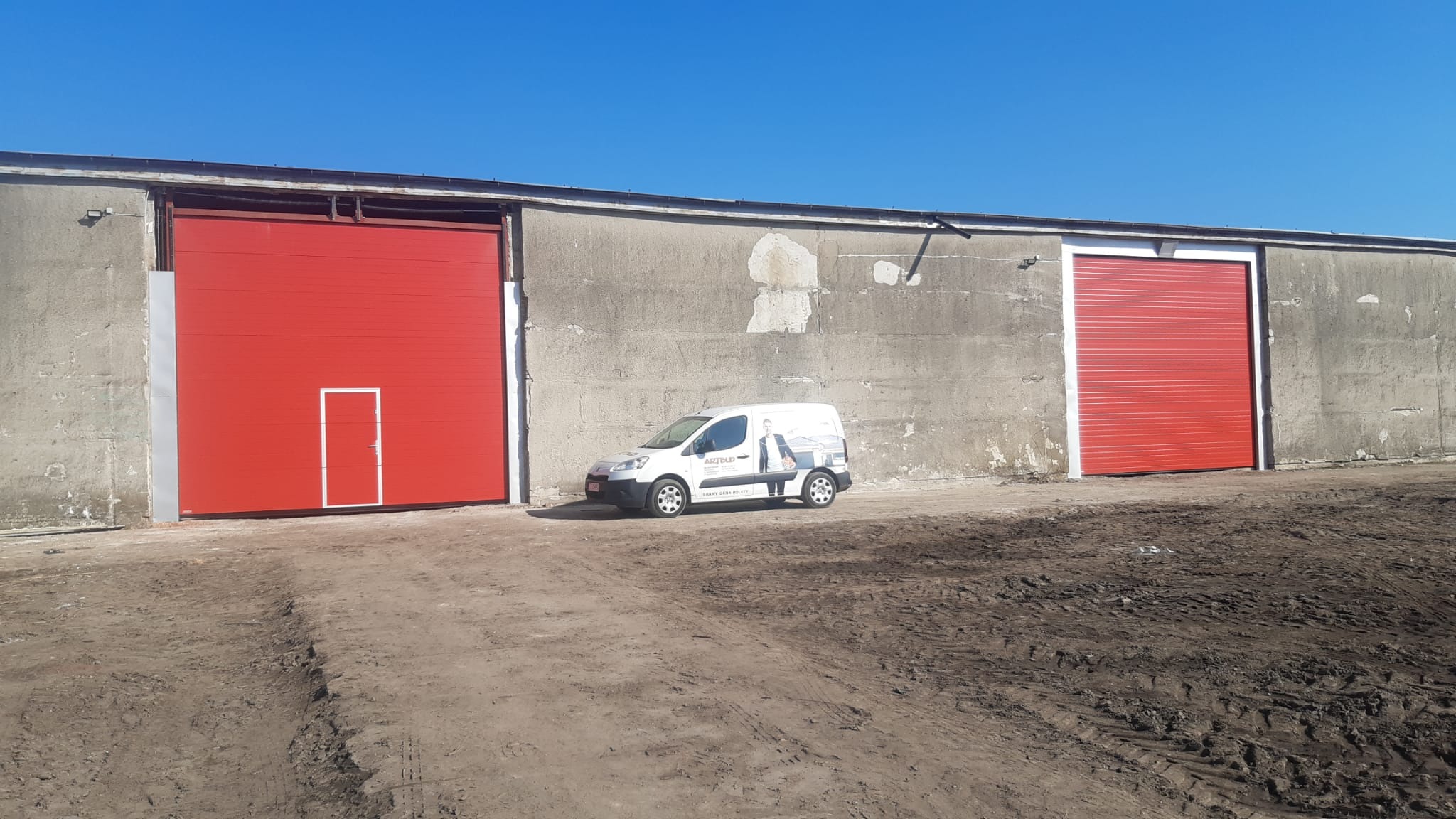 Czerwone segmentowe bramy garażowe w szarym betonowym budynku przemysłowym, biały samochód dostawczy z reklamą firmy parkuje na ziemi.