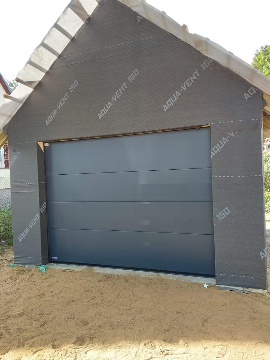 Szara brama garażowa segmentowa, widok z zewnątrz podczas montażu, ściany budynku pokryte membraną izolacyjną z nadrukiem Aqua-Vent 150, piasek na podłożu.